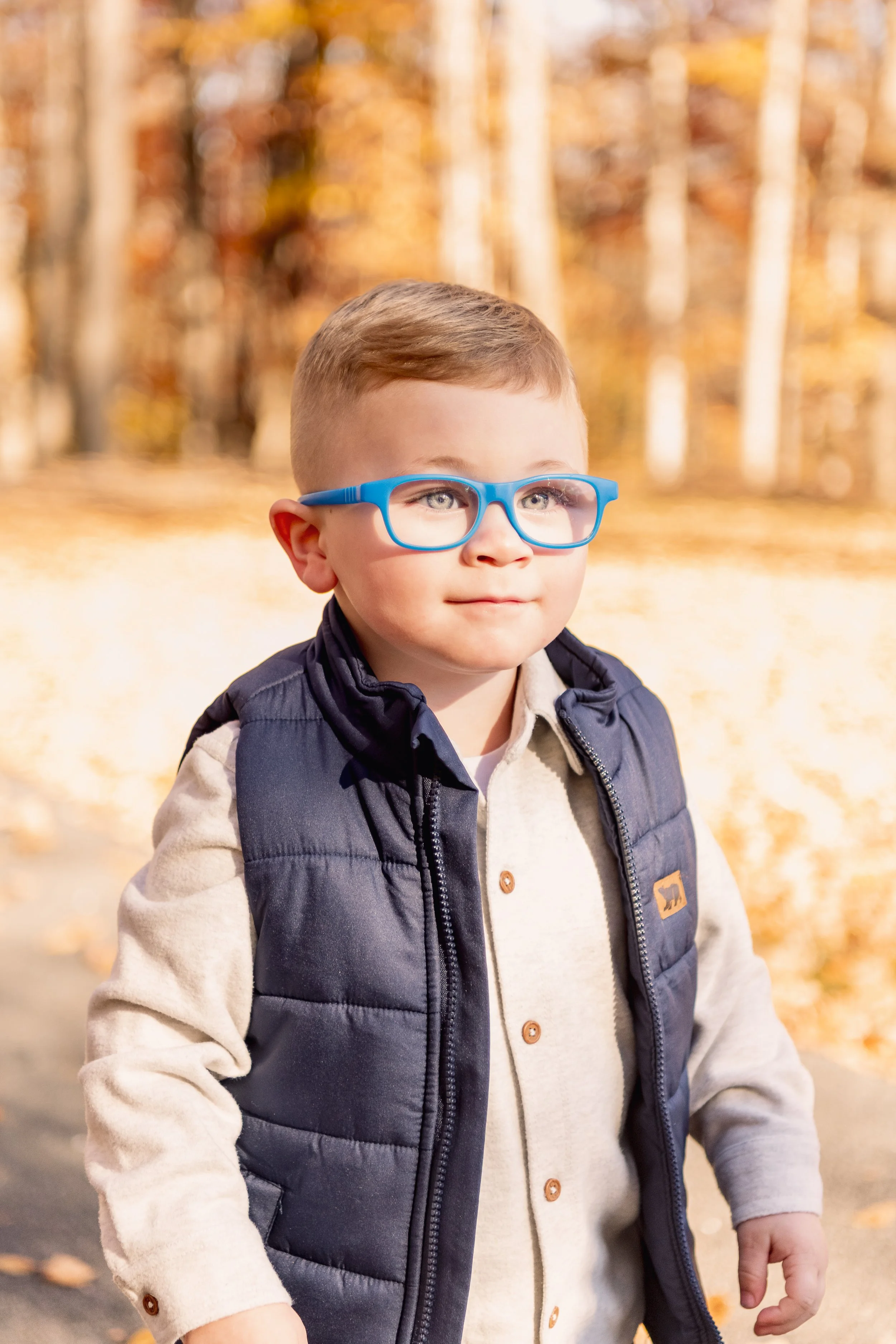 Young boy with light brown hair and glasses wearing a puffer vest and khaki shirt standing outdoors in autumn