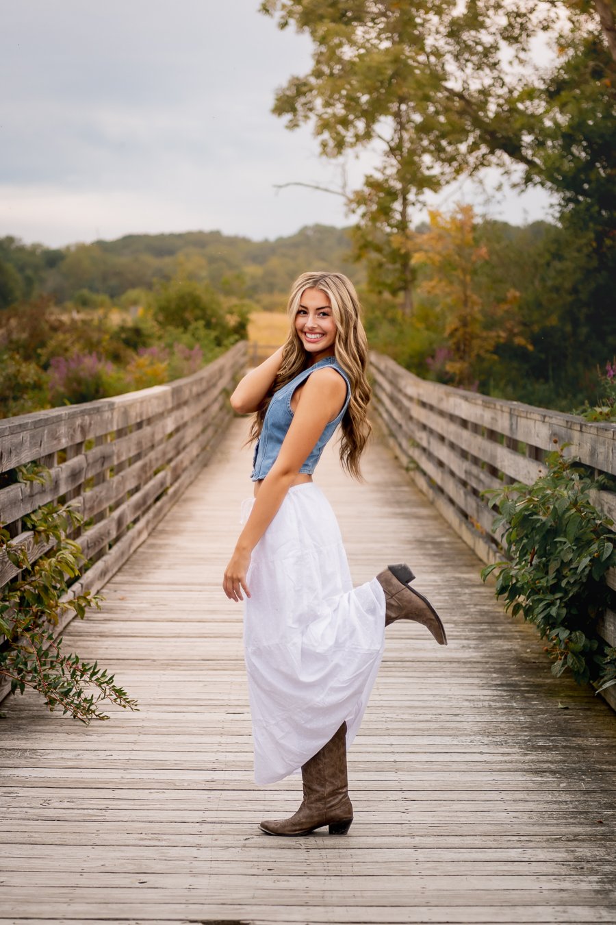 A young woman stands on a wooden bridge outdoors, smiling and lifting her right leg. She has long wavy hair and is wearing a sleeveless denim top, a white skirt, and brown cowboy boots. The background shows trees with fall foliage and a cloudy sky.