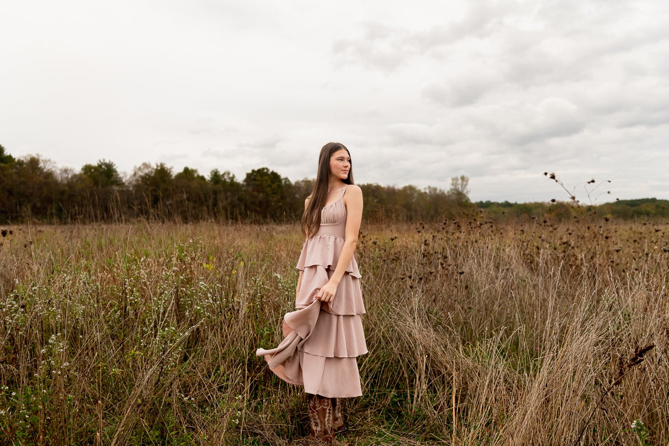 A woman in a flowing beige dress standing in a field of tall grass and wildflowers under a cloudy sky.
