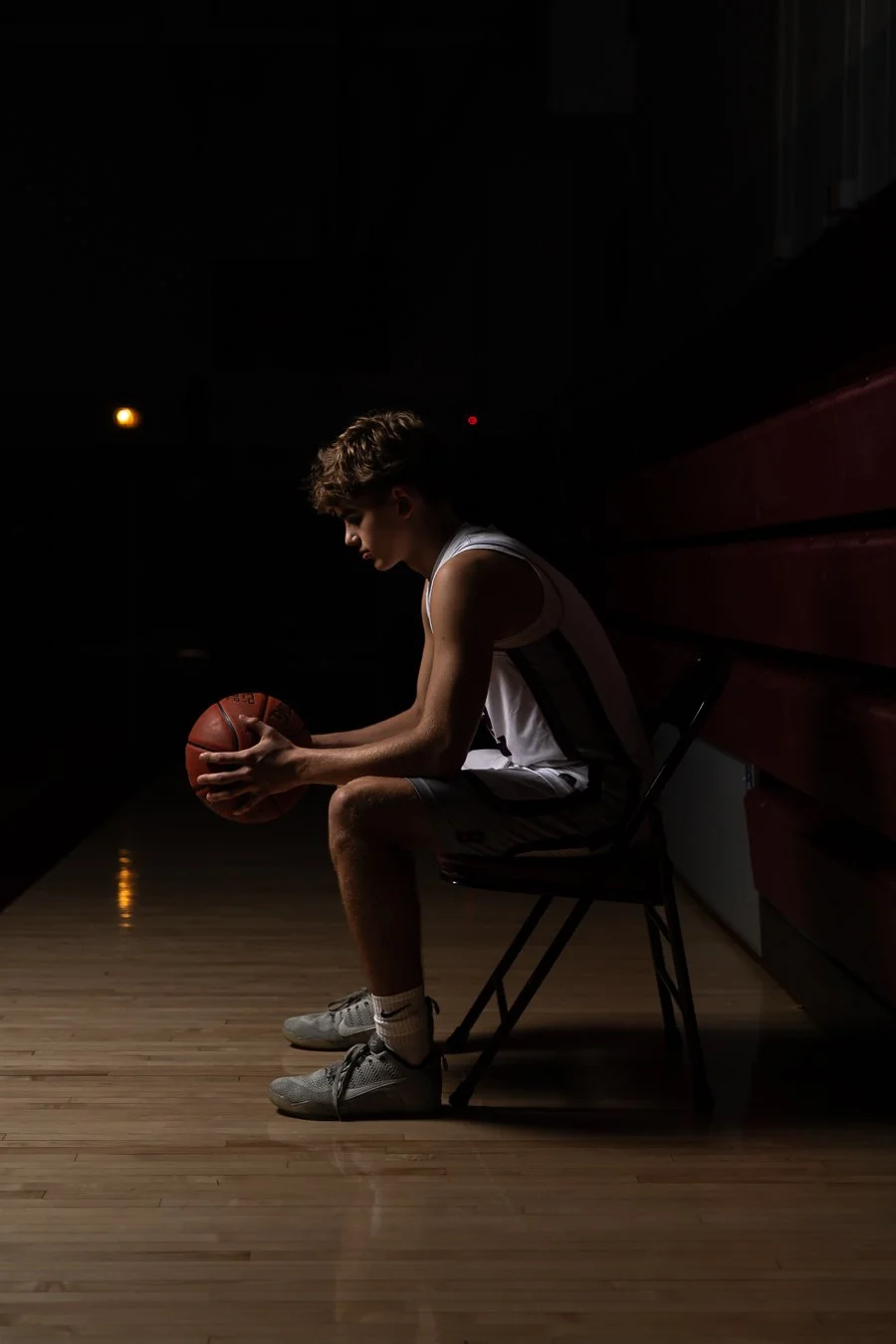 A young male basketball player sitting on a bench in a dark gym, holding a basketball, with a contemplative expression.
