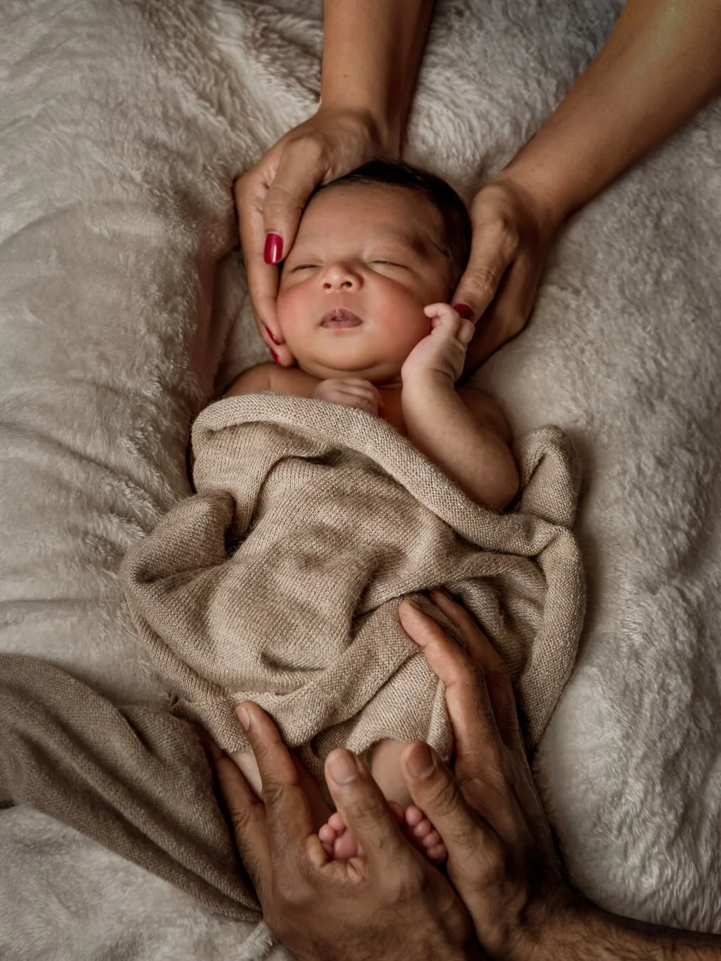 A newborn baby peacefully sleeping on a soft, beige blanket, with hands gently supporting the baby's head and feet.