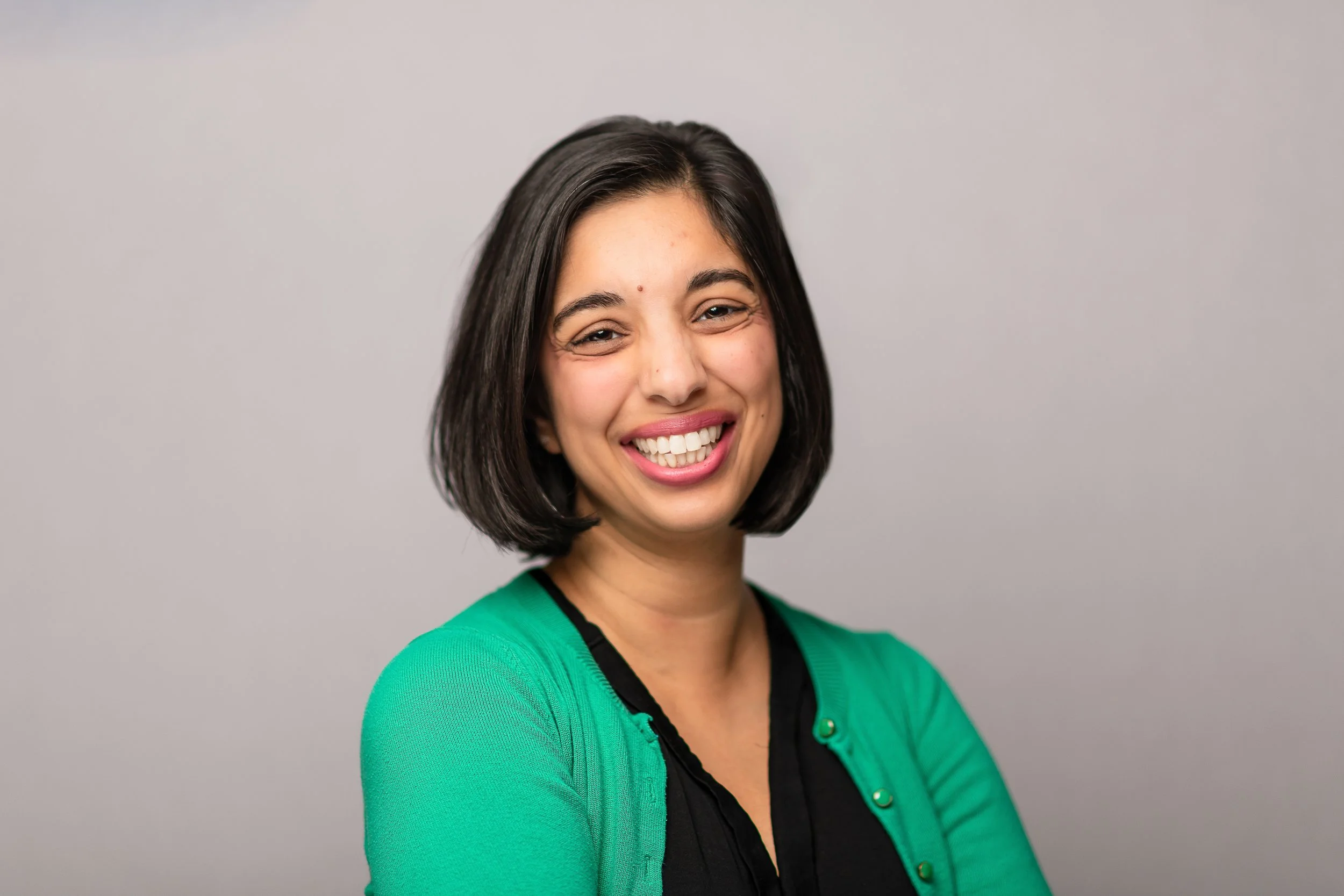 A woman with short black hair smiling at the camera, wearing a green cardigan over a black top, standing against a plain light gray background.
