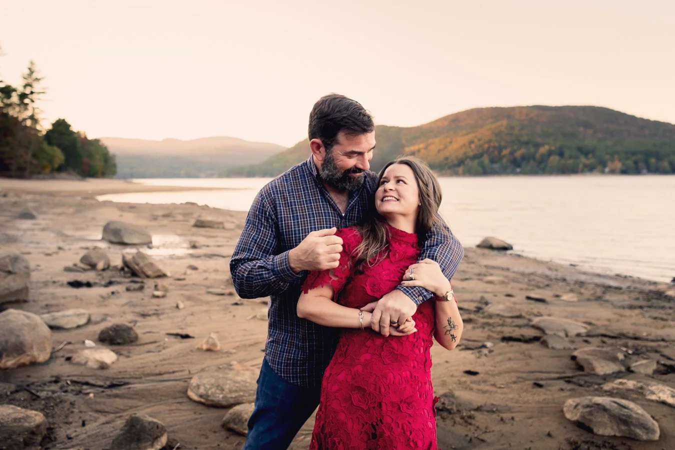 A couple standing on a rocky beach with a lake and mountains in the background during sunset, embracing and smiling at each other.