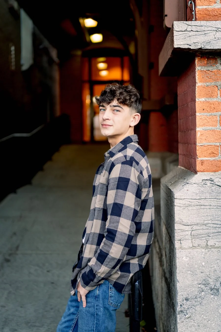 A young man with curly dark hair and light skin is leaning against a brick and stone wall inside a building, wearing a checkered shirt and blue jeans, looking towards the camera.