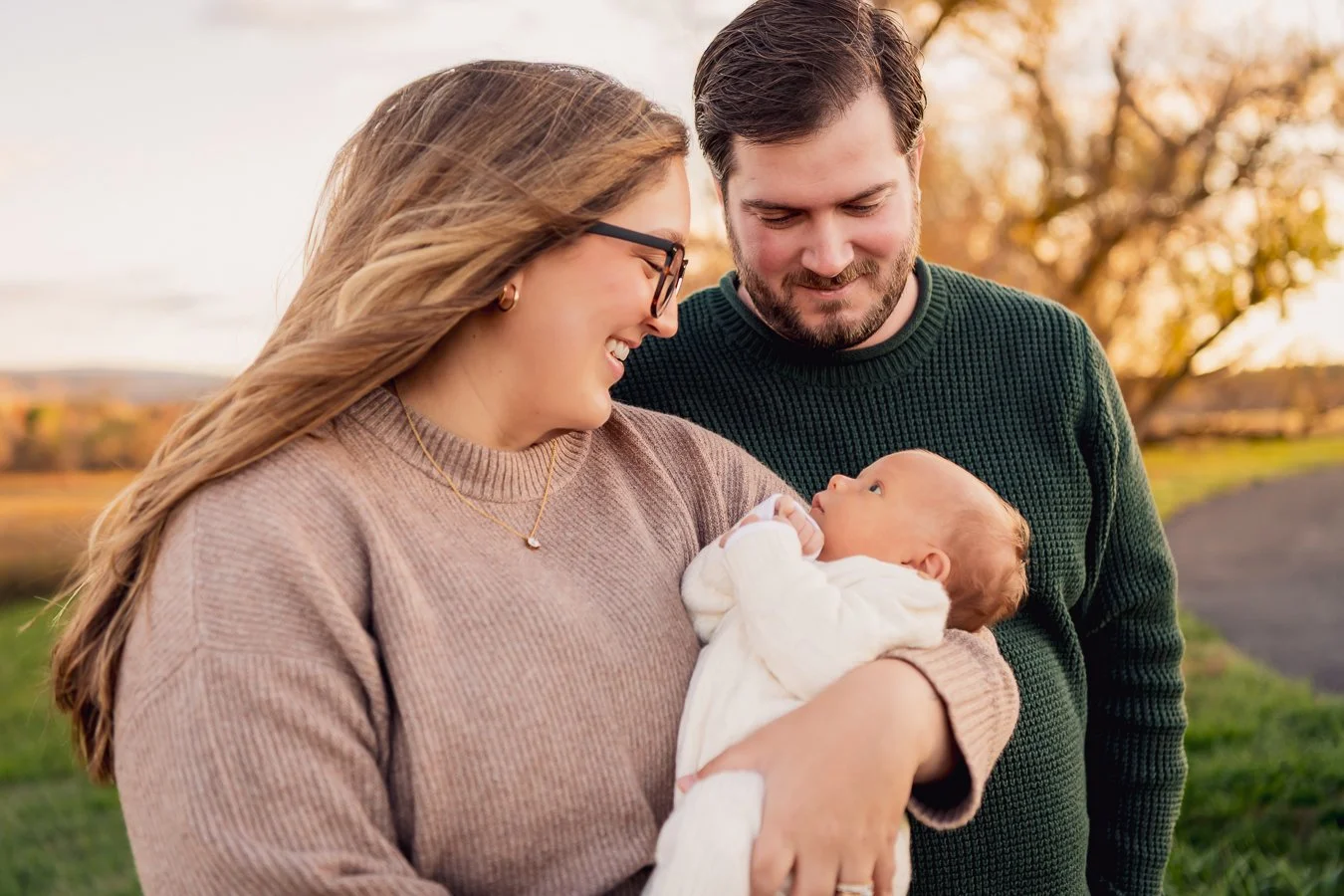 A happy couple holding a newborn baby outdoors during sunset, smiling at each other and the baby, with trees and a grassy field in the background.