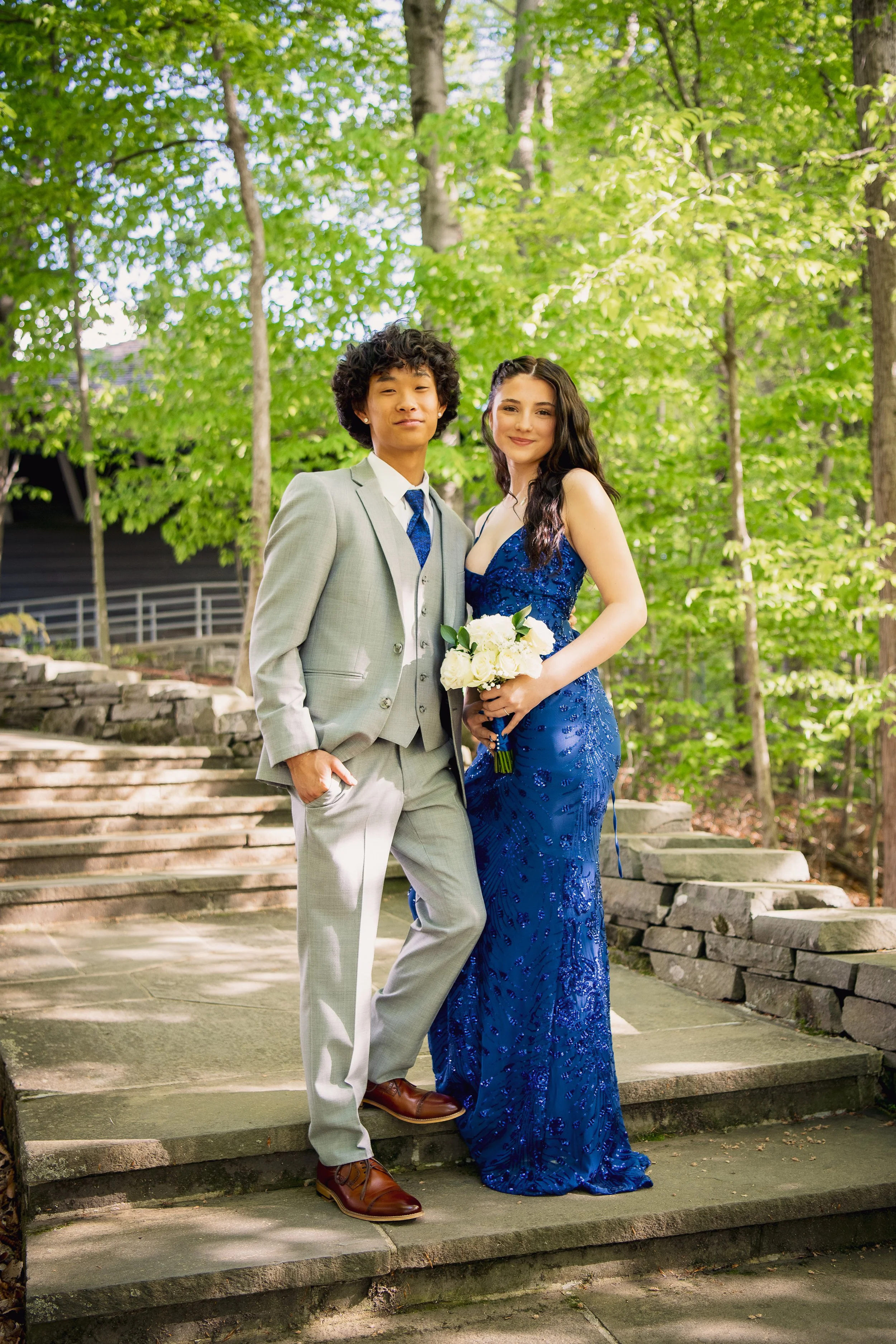 A young man in a light gray suit with a blue tie and a young woman in a long, royal blue gown holding a bouquet of white roses, standing on stone steps in a wooded park during daytime.