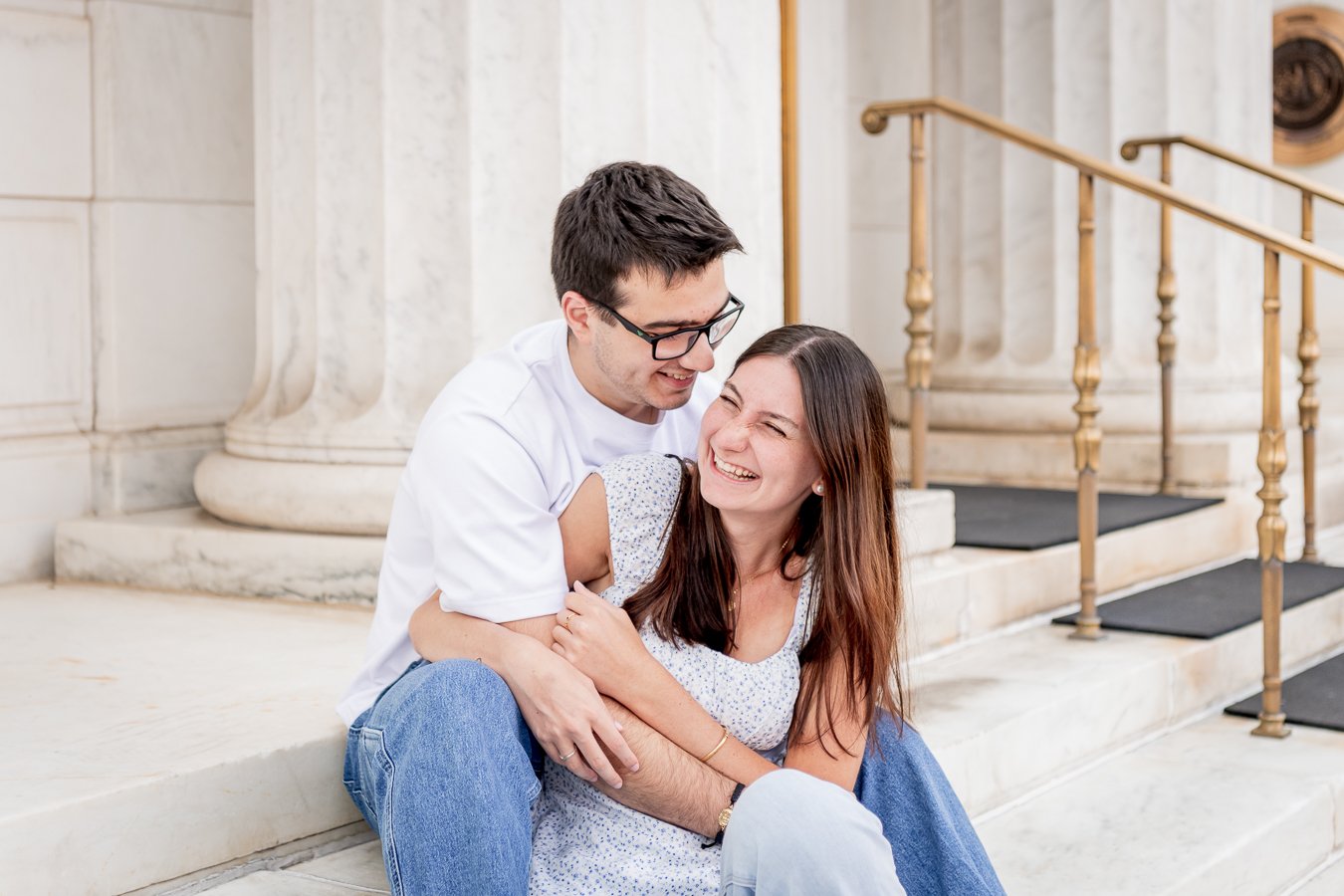 A happy couple sitting on steps outside, laughing and embracing each other near a building with marble columns.