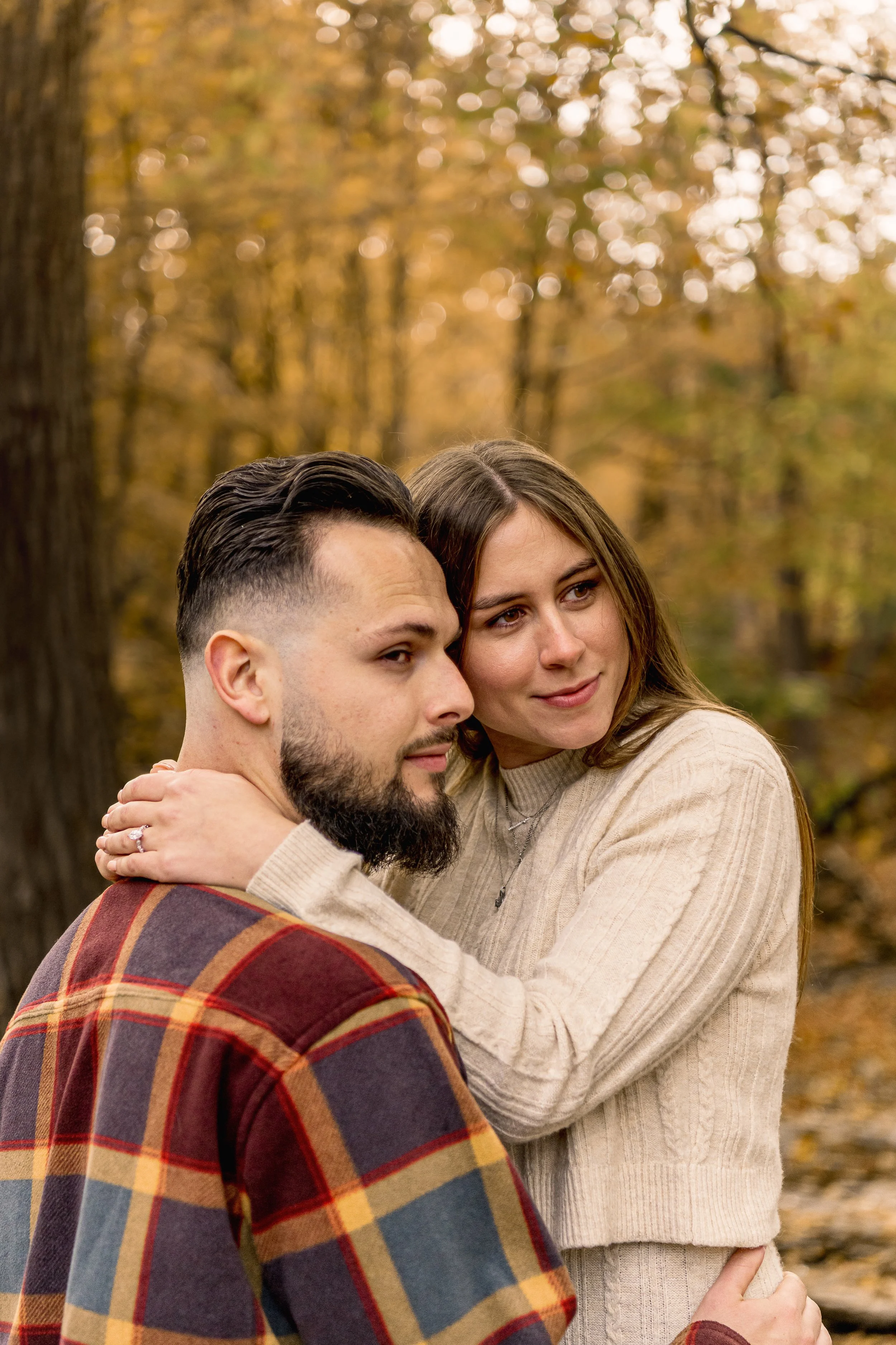 A couple embracing outdoors during autumn, with trees and fall foliage in the background.