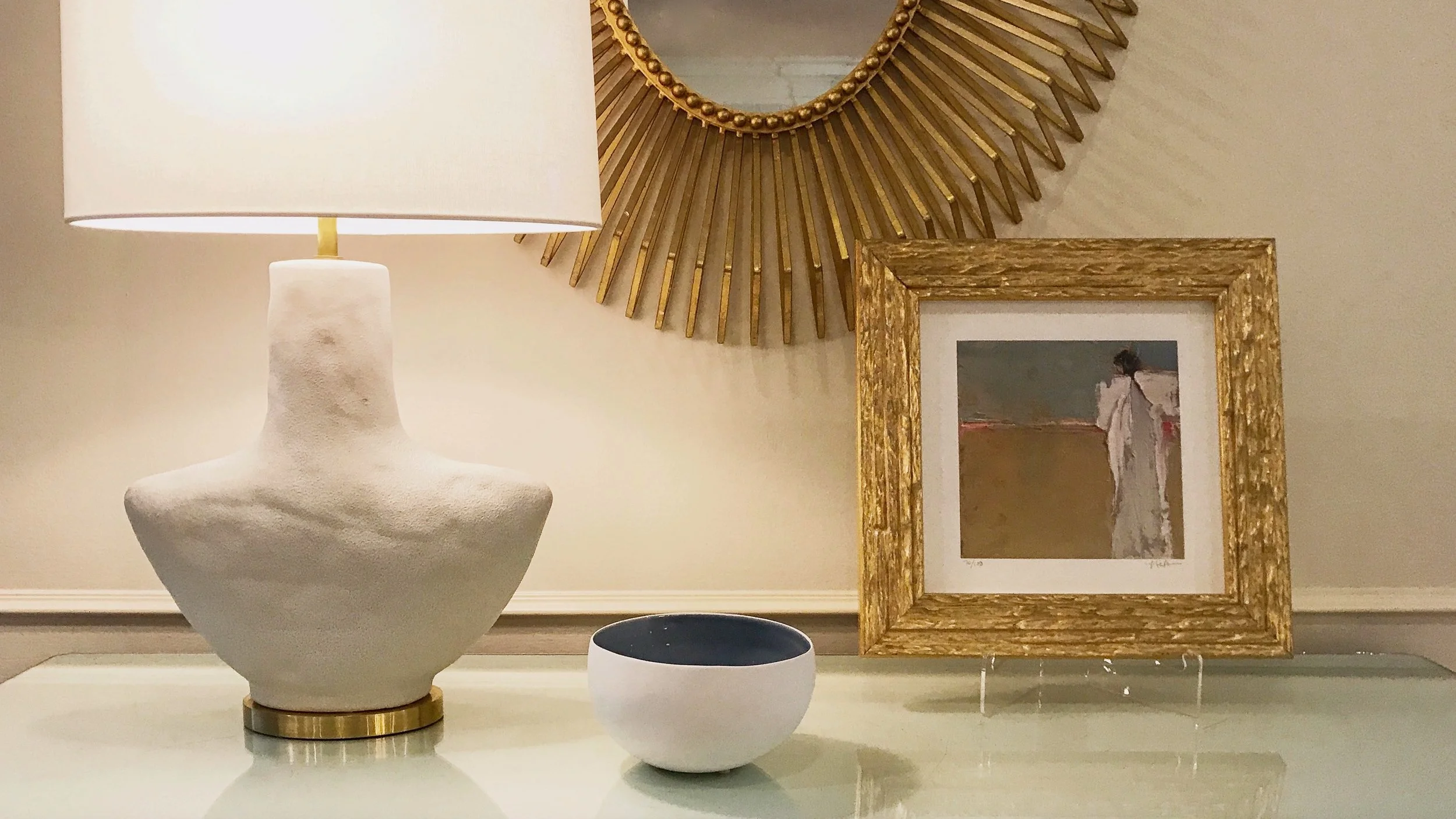A beige table with a white textured table lamp, a small black and white bowl, a framed painting of a woman in a white dress, and a large round mirror with gold rays on the wall.
