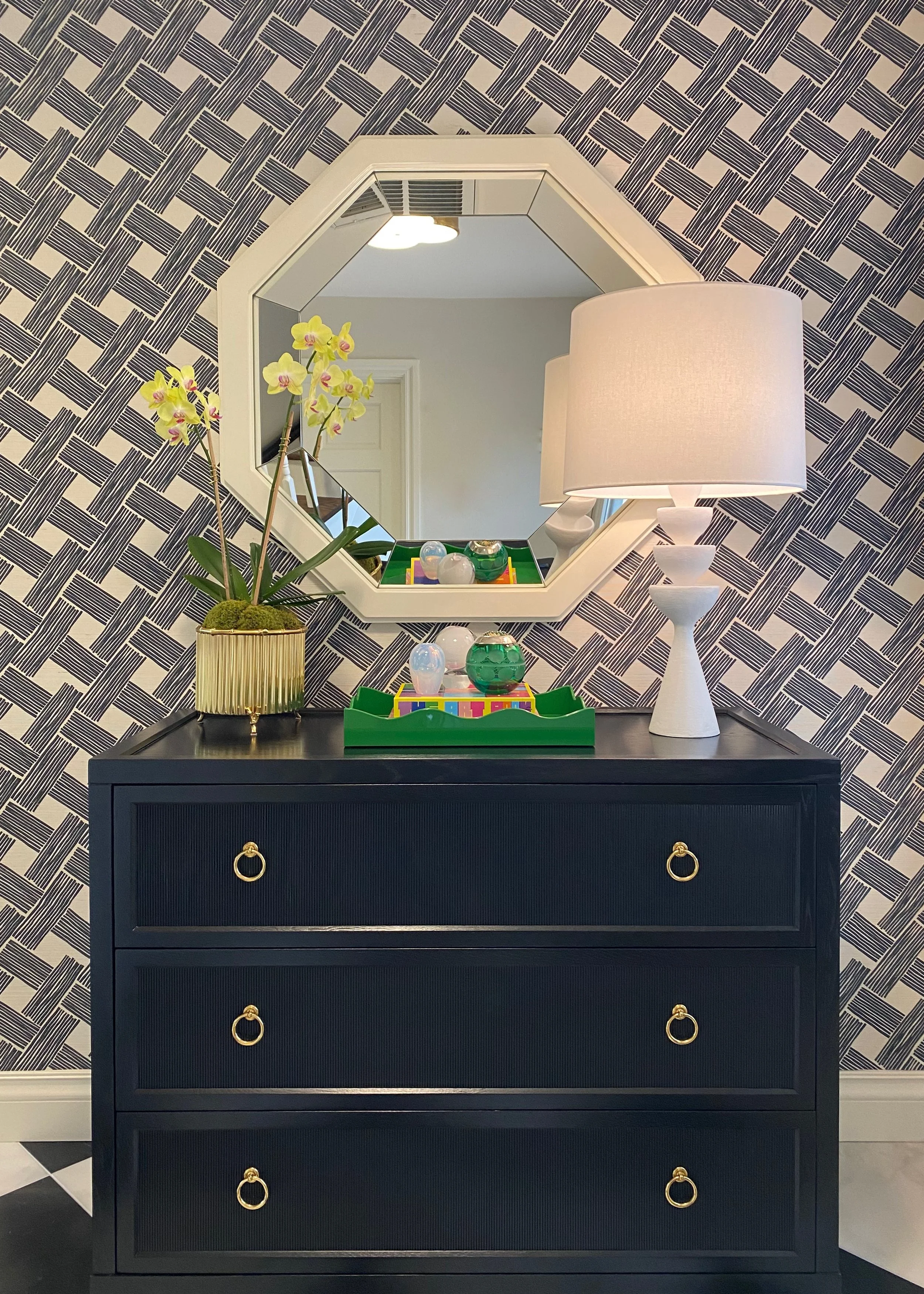 A black dresser with three drawers and gold ring handles, topped with a white table lamp, a yellow orchid in a gold vase, and a green tray with decorative glass spheres, all reflected in an octagonal mirror on a wallpapered wall with a geometric black and white pattern.