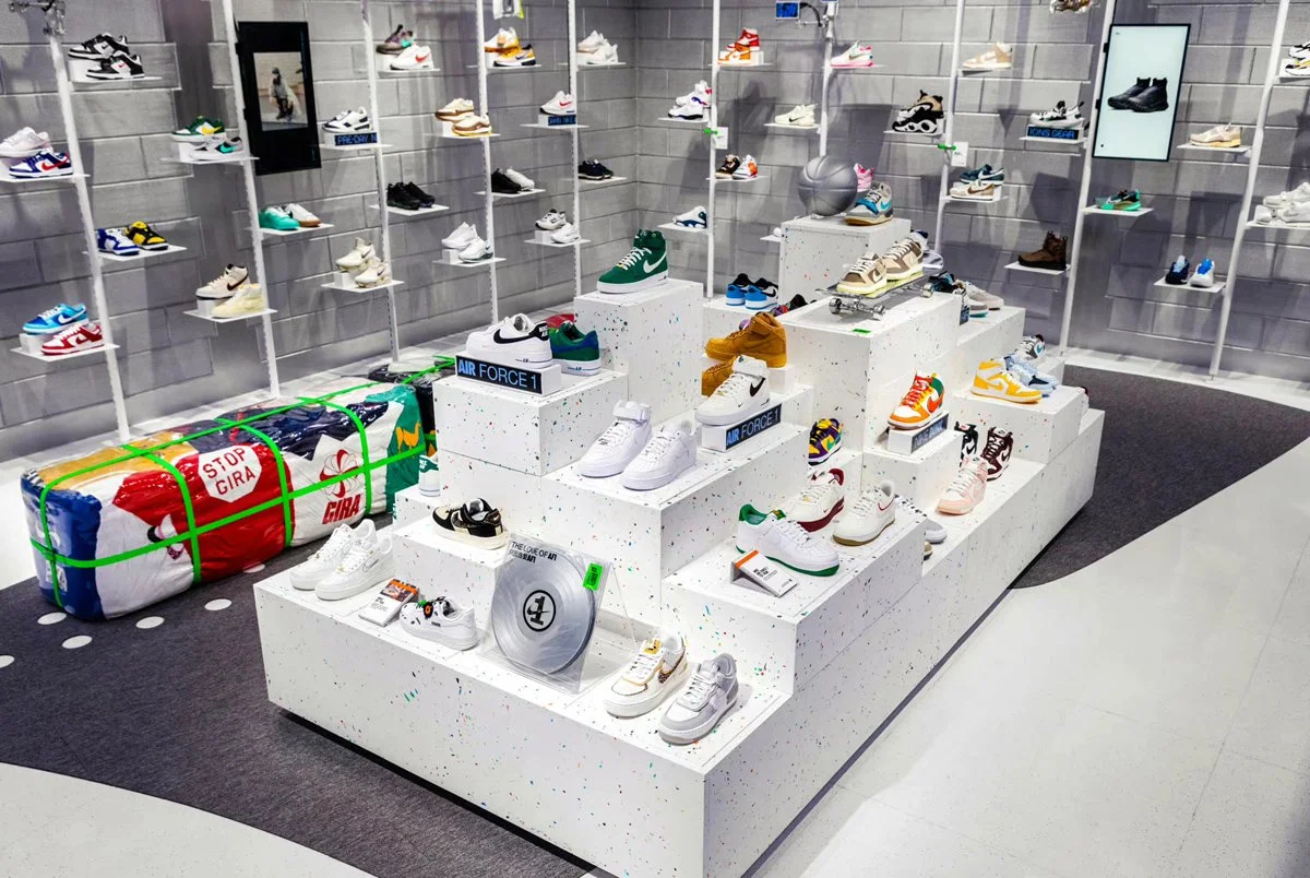 Display of various sneakers on tiered white platforms in a retail store, with shelves of shoes on a gray wall in the background.