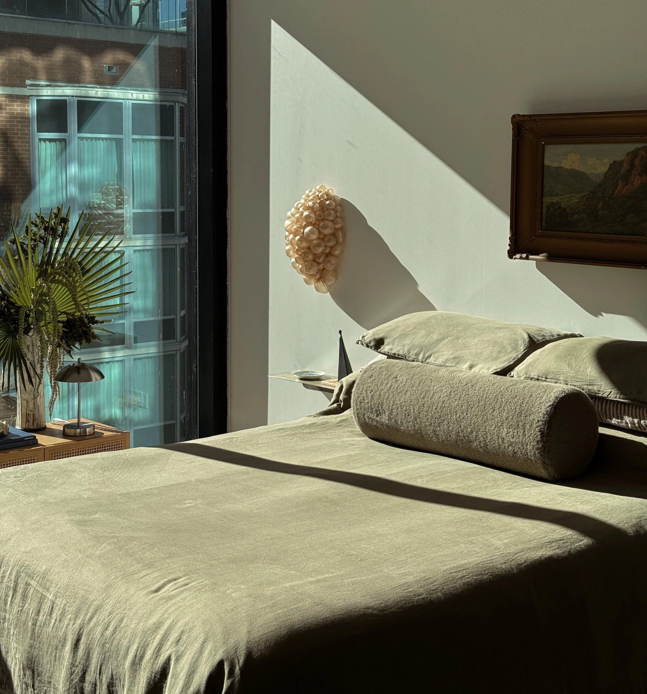 A cozy bedroom corner with a bed covered in green fabric, two matching pillows, and a bolster pillow. Sunlight through a large window creates shadows on the bed and wall. A small side table holds a plant, a desk lamp, and some books. A decorative wall piece made of shells or beads is mounted on the wall, and there is a landscape painting in a wooden frame.