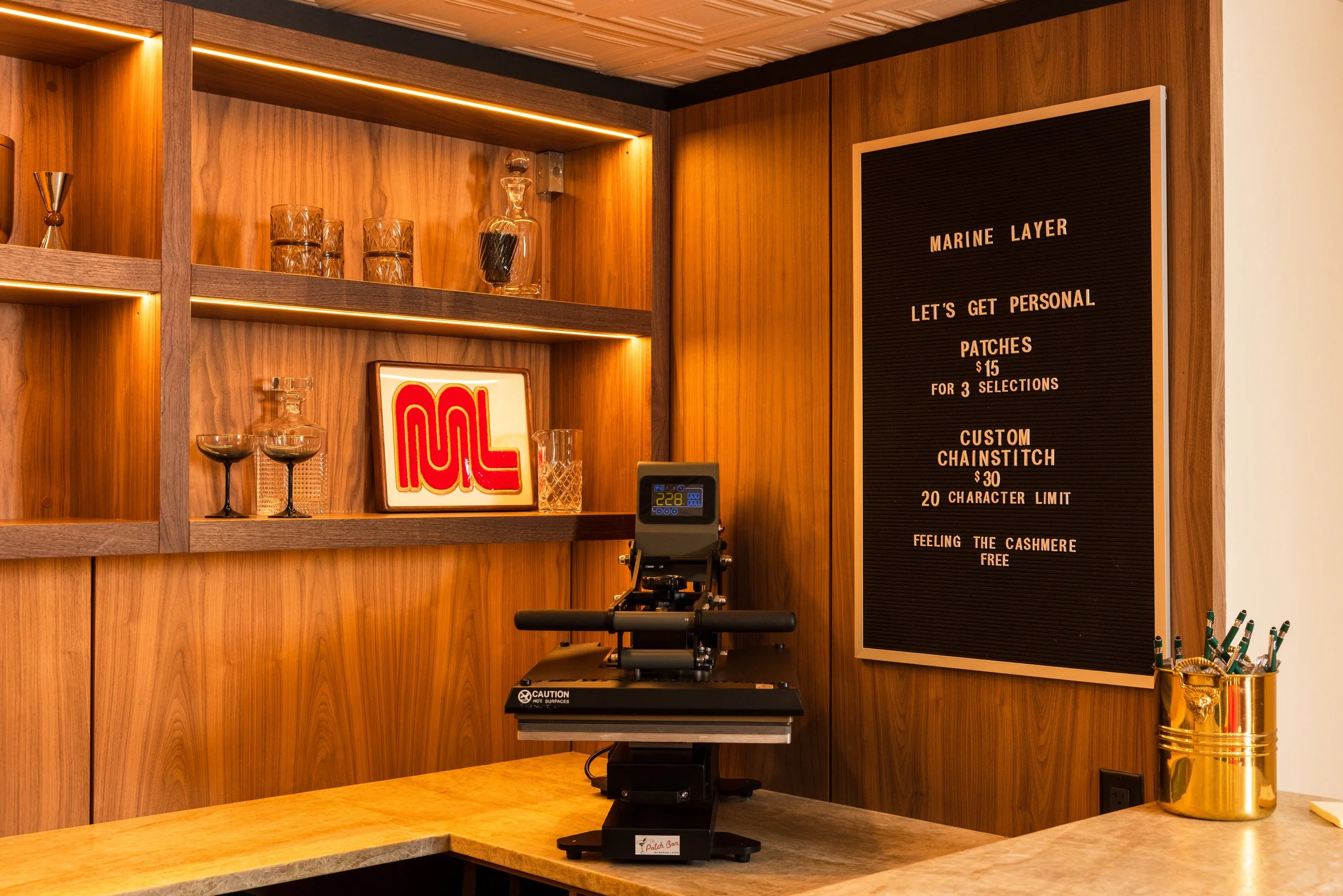 A retail space with wooden shelving and a black menu board displaying prices for patches and chainstitch, a digital scale on a countertop, glassware and a framed artwork on the shelves, and a gold pen holder on the counter.