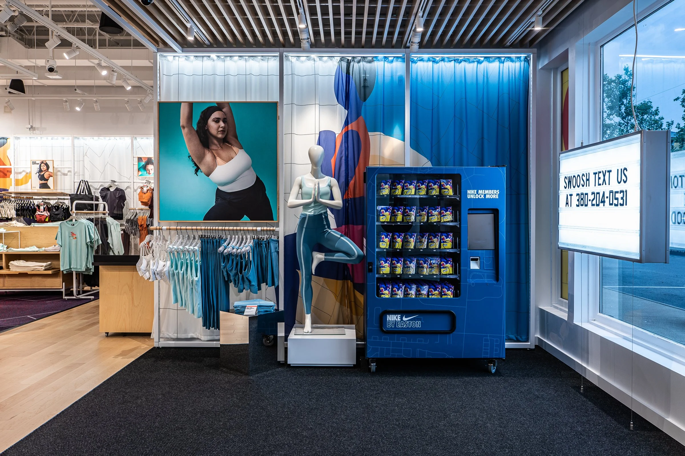 Sportswear display inside a retail store with a mannequin performing a yoga pose, promotional posters, clothing racks, a vending machine, and a bright sign with store contact information.