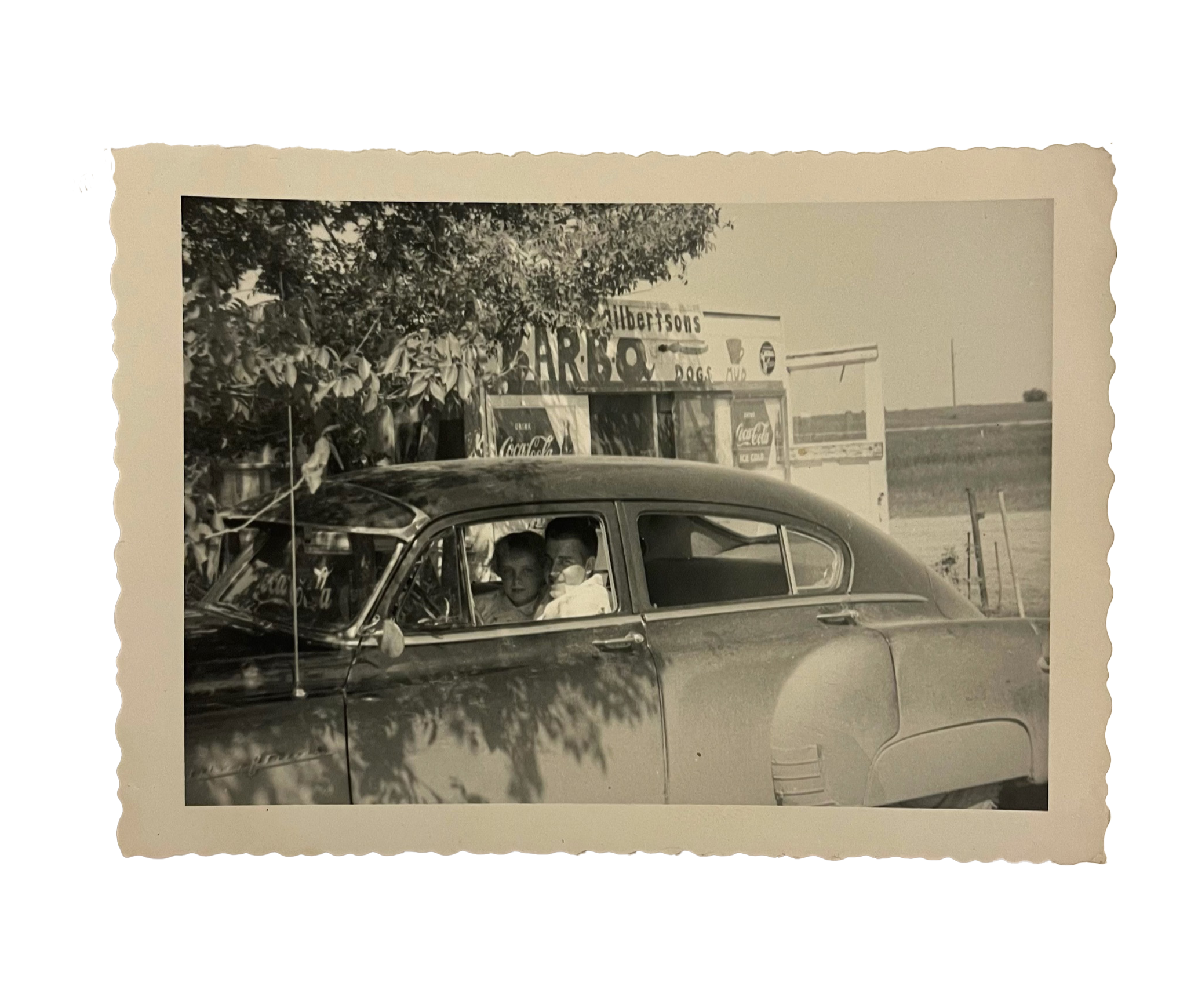 Black and white photo of a vintage car parked in front of a food stand with two children sitting inside the car, in a rural area.