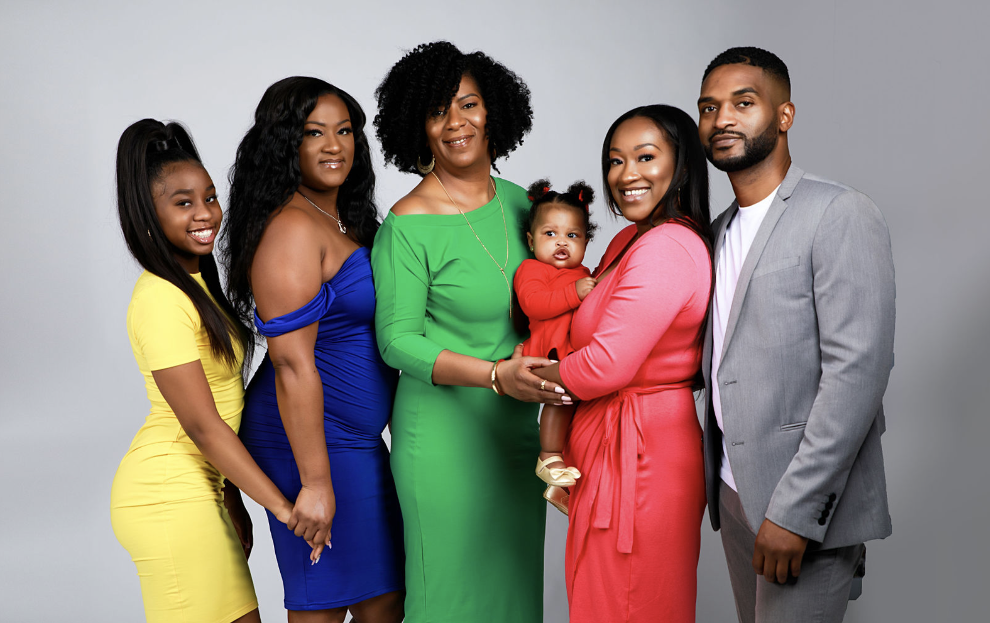 Studio family portrait of a grandmother with her adult children and baby granddaughter standing together, photographed to highlight generational connection.