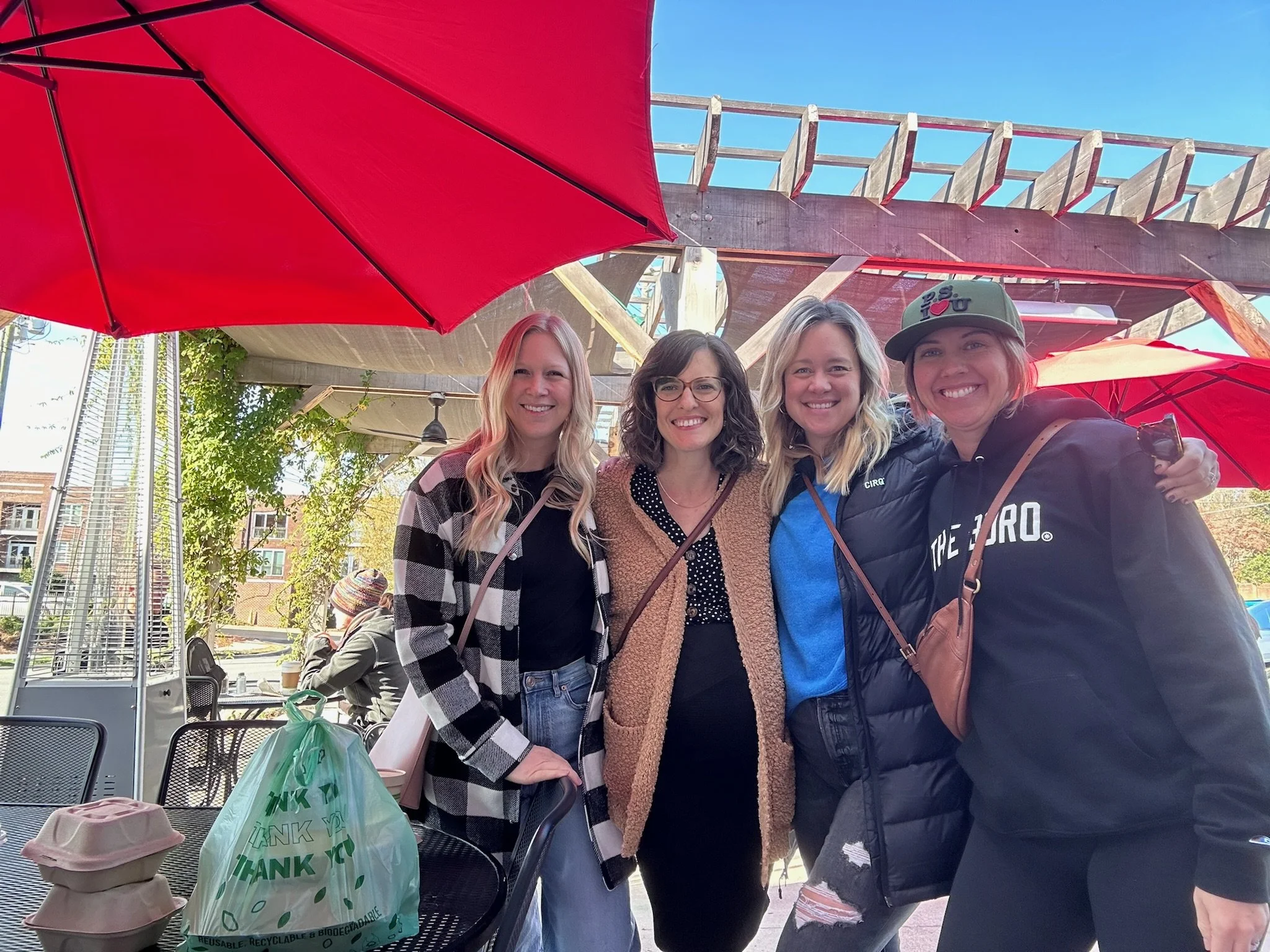 Four women smiling and posing together outdoors under a large red umbrella at a cafe or restaurant. They are dressed casually, with various hair colors and styles. In the foreground, there are takeaway coffee cups, a green plastic bag, and some food containers on a table.