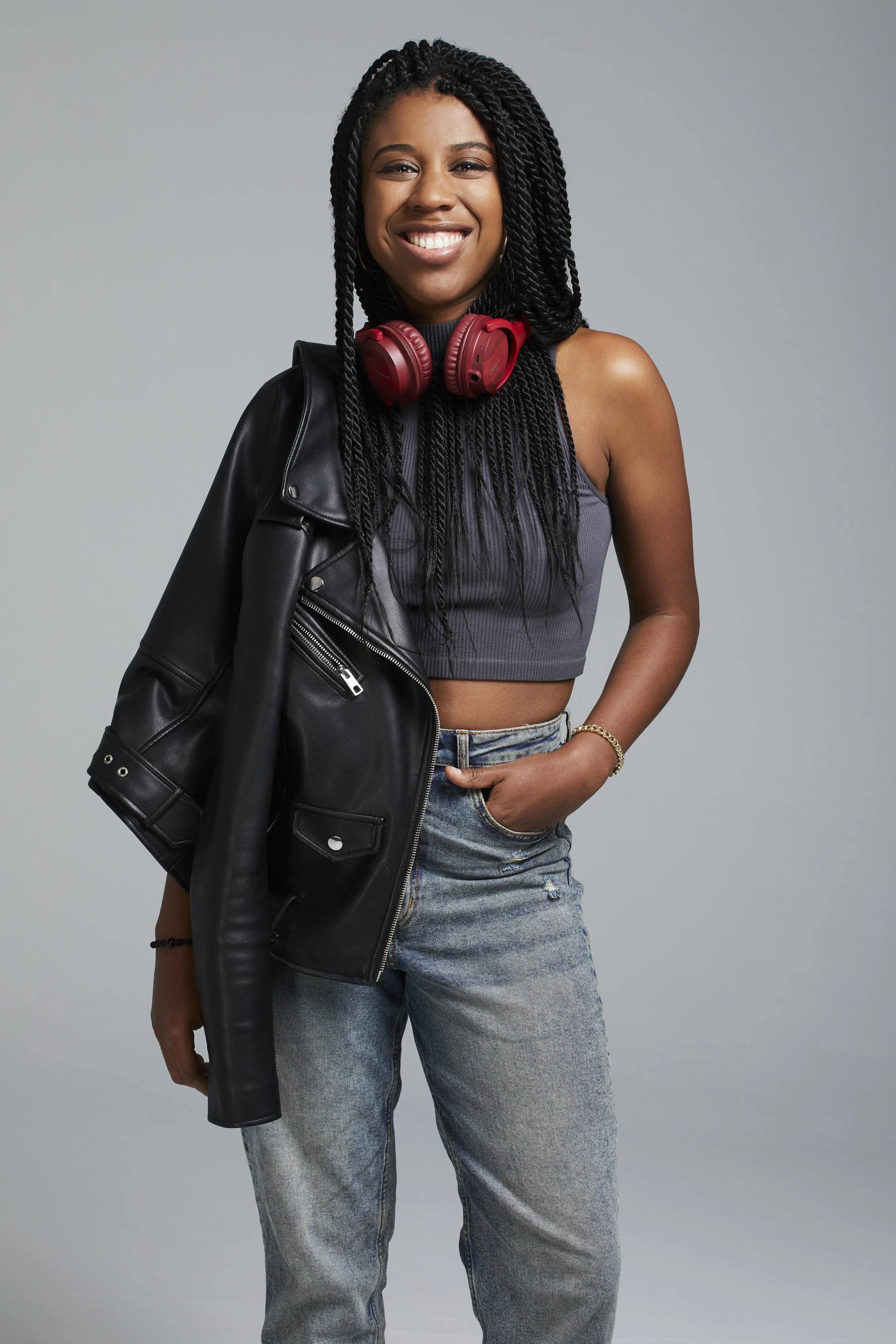 A young woman with braided hair smiling, wearing a gray crop top, light blue jeans, a black leather jacket slung over her shoulder, and red headphones around her neck.