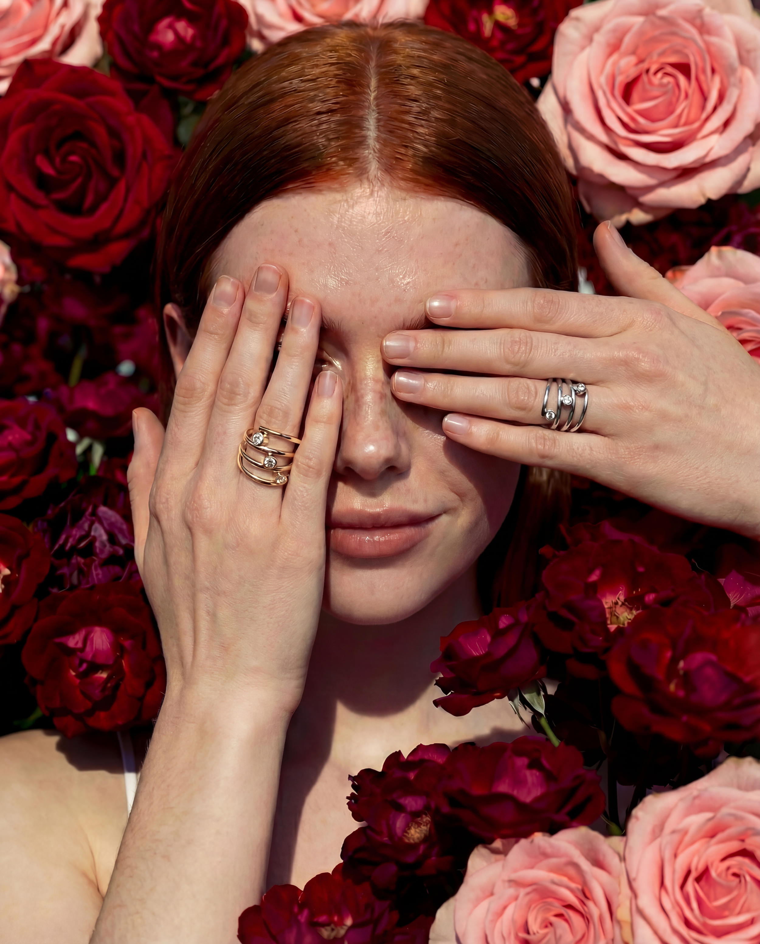 Red haired model covers both eyes with hands while showing two diamond rings, one in gold and one in silver.