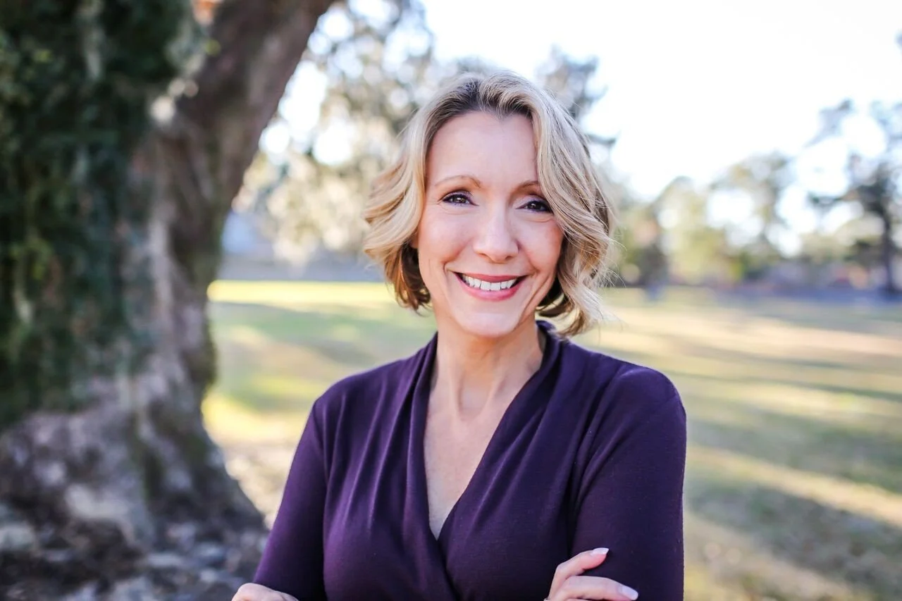 A smiling woman with blonde hair in a purple top standing outdoors near a tree in a park.