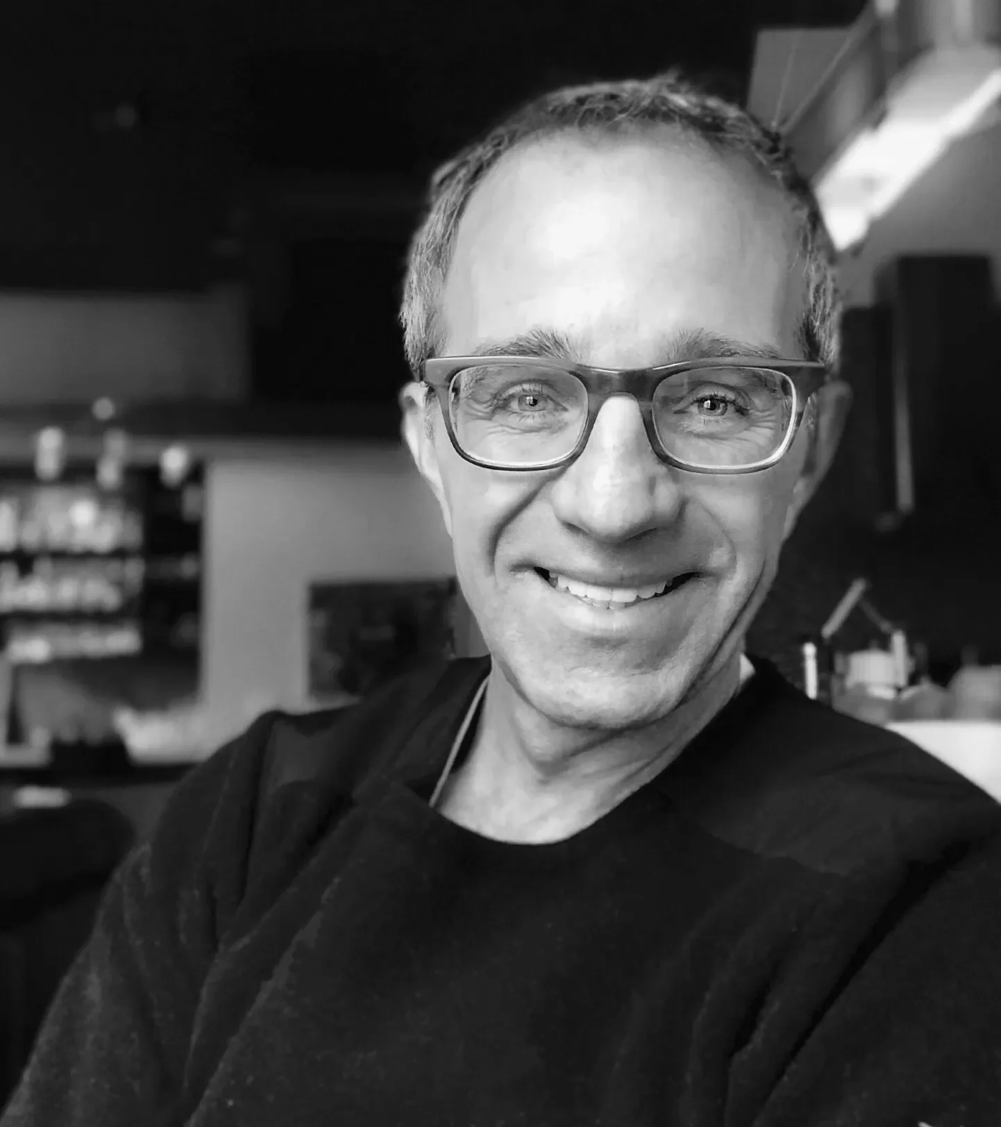 A smiling man with glasses and short hair taking a selfie in a room with shelves in the background.