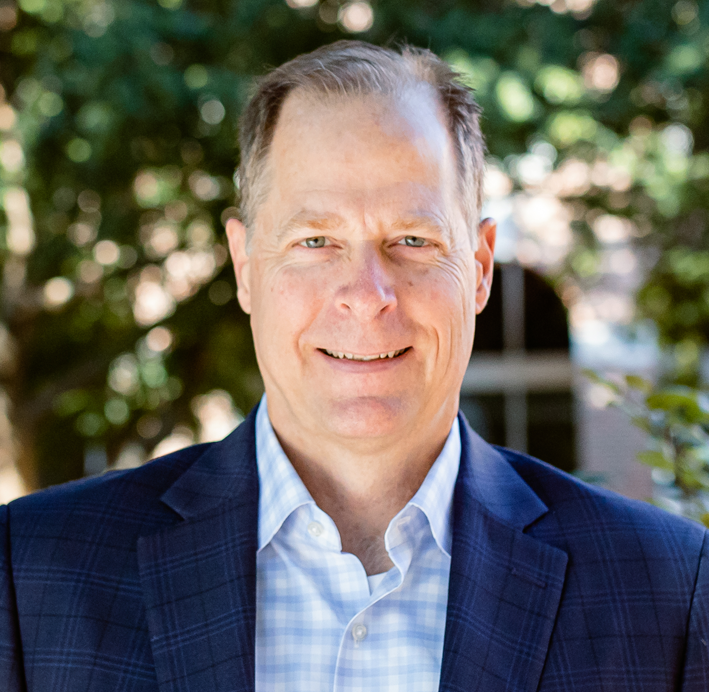 A middle-aged man with light-colored hair smiling outdoors, wearing a navy blue blazer and a light blue checkered shirt, with trees and blurred background.