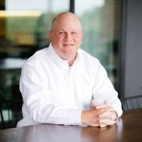A man with glasses and a shaved head wearing a white shirt, sitting at a table in an office or conference room.