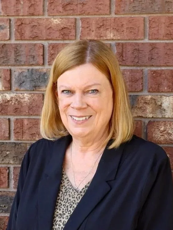 A woman with light skin, shoulder-length blonde hair, and blue eyes standing in front of a brick wall. She is smiling and wearing a black blazer over a leopard print top.