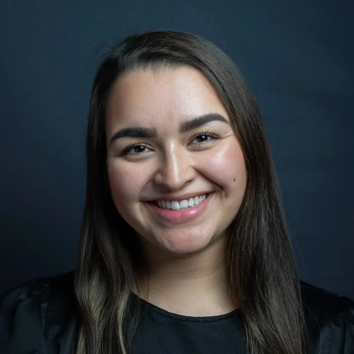 A young woman with long brown hair, smiling, wearing a black top, against a dark background.