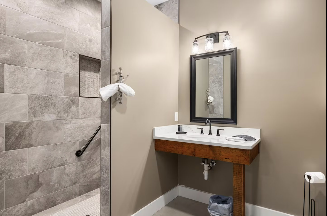 Bathroom with beige painted walls, a walk-in shower with tiled walls, a black-framed mirror above a white marble sink, and a trash can underneath. There is a towel hanging on the shower pipe, and a towel and toiletries on the sink.
