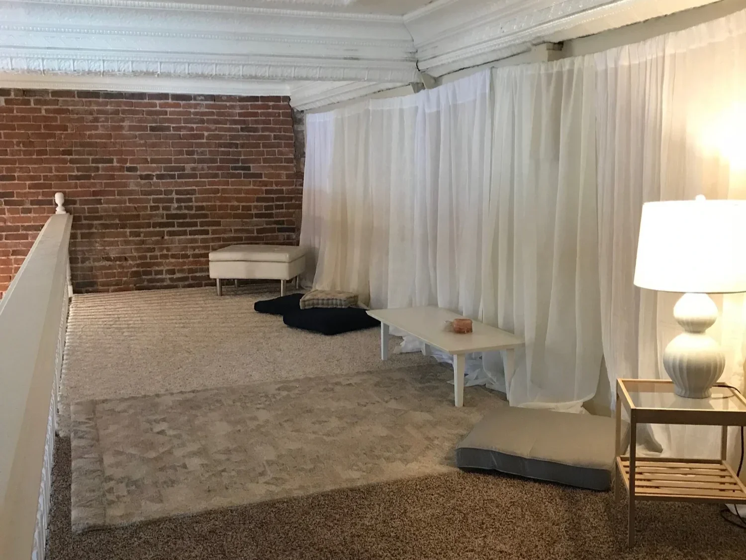 Living space with exposed brick wall, white curtains, beige carpet, a white table, a lamp on side table, and pets bed and cushions on the floor.