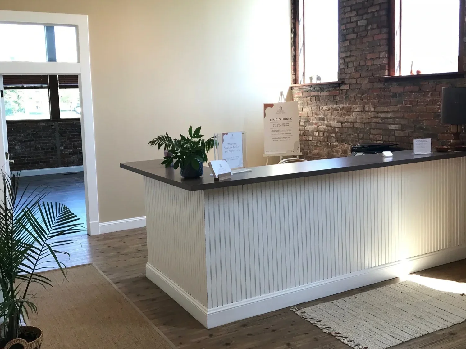 Reception desk with a small potted plant, informational signs, brick wall behind, and large windows letting in natural light.
