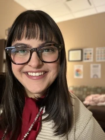 Close-up of a woman with dark hair, wearing glasses, smiling, in an indoor setting with framed pictures on the wall behind her.