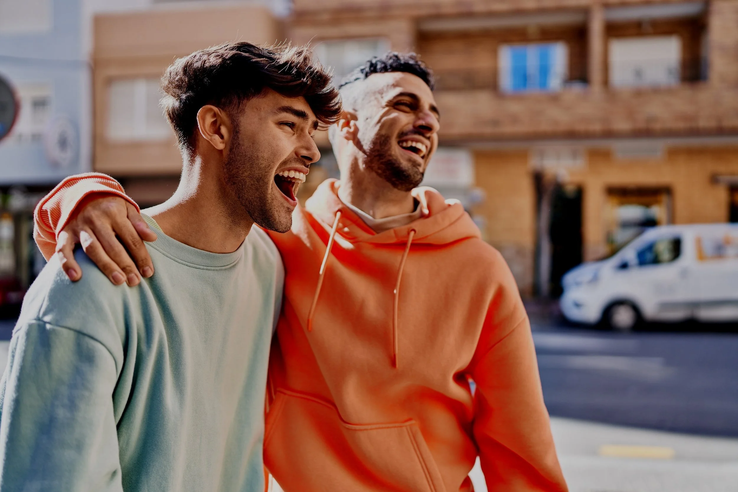 Two young men walking together outdoors, smiling and laughing, with one man putting his arm around the other.