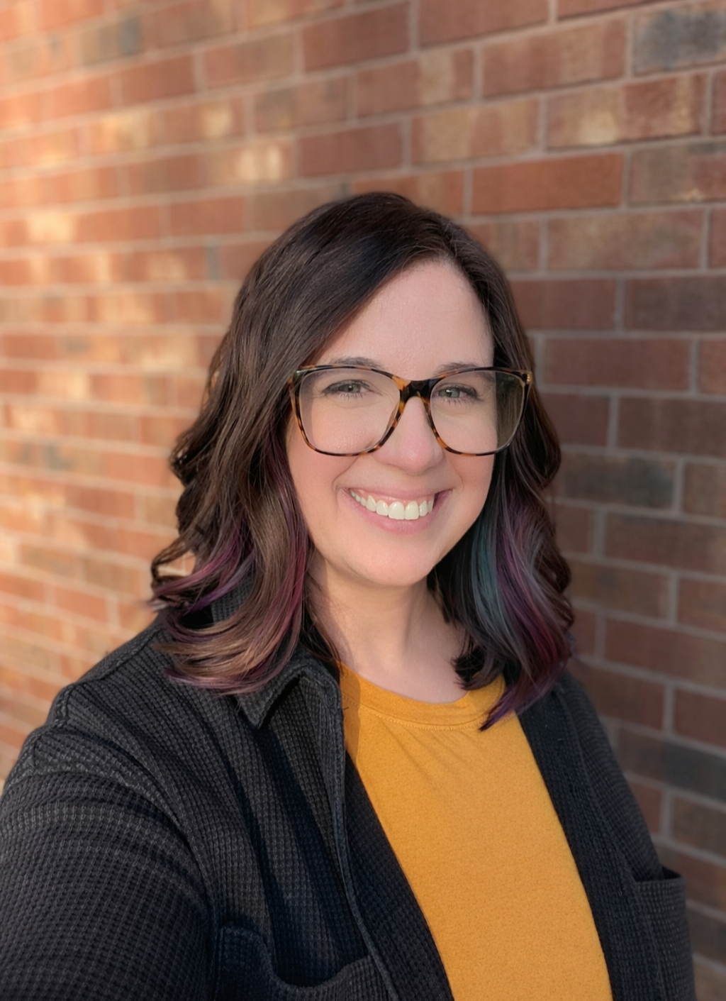 A woman with shoulder-length wavy dark hair tinted with purple and blue highlights, wearing tortoiseshell glasses, a yellow shirt, and a black textured jacket, smiling in front of a brick wall.