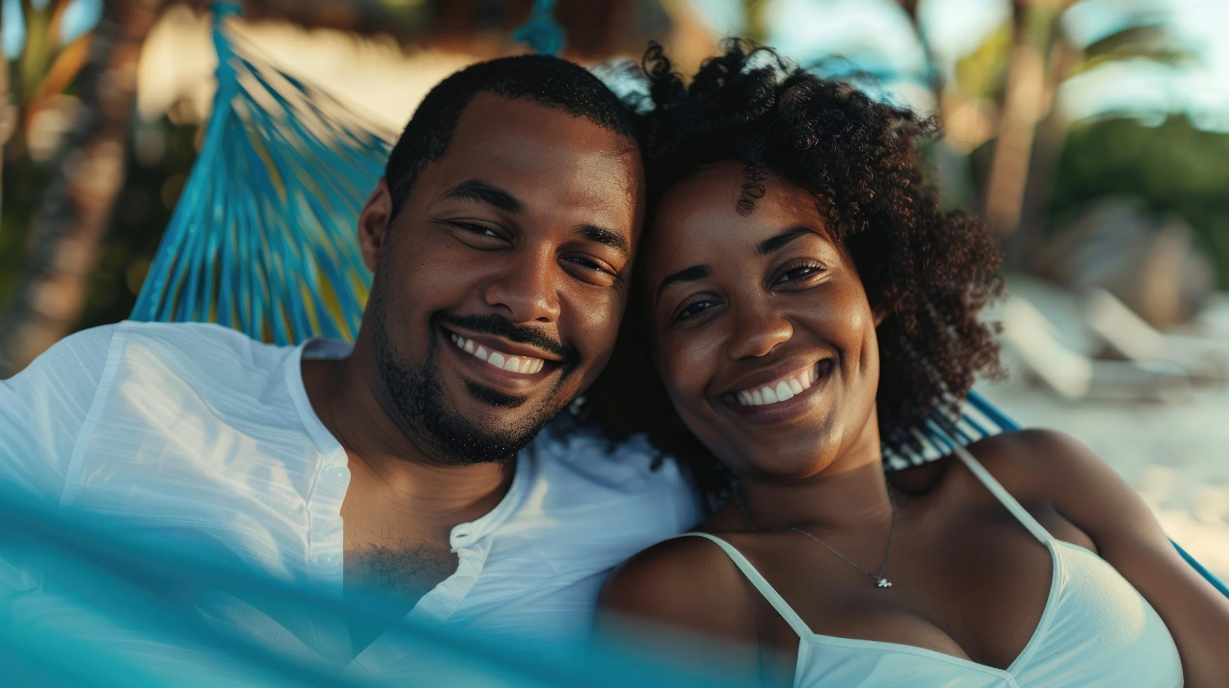 A smiling couple taking a selfie at the beach, relaxing on a hammock with palm trees in the background.
