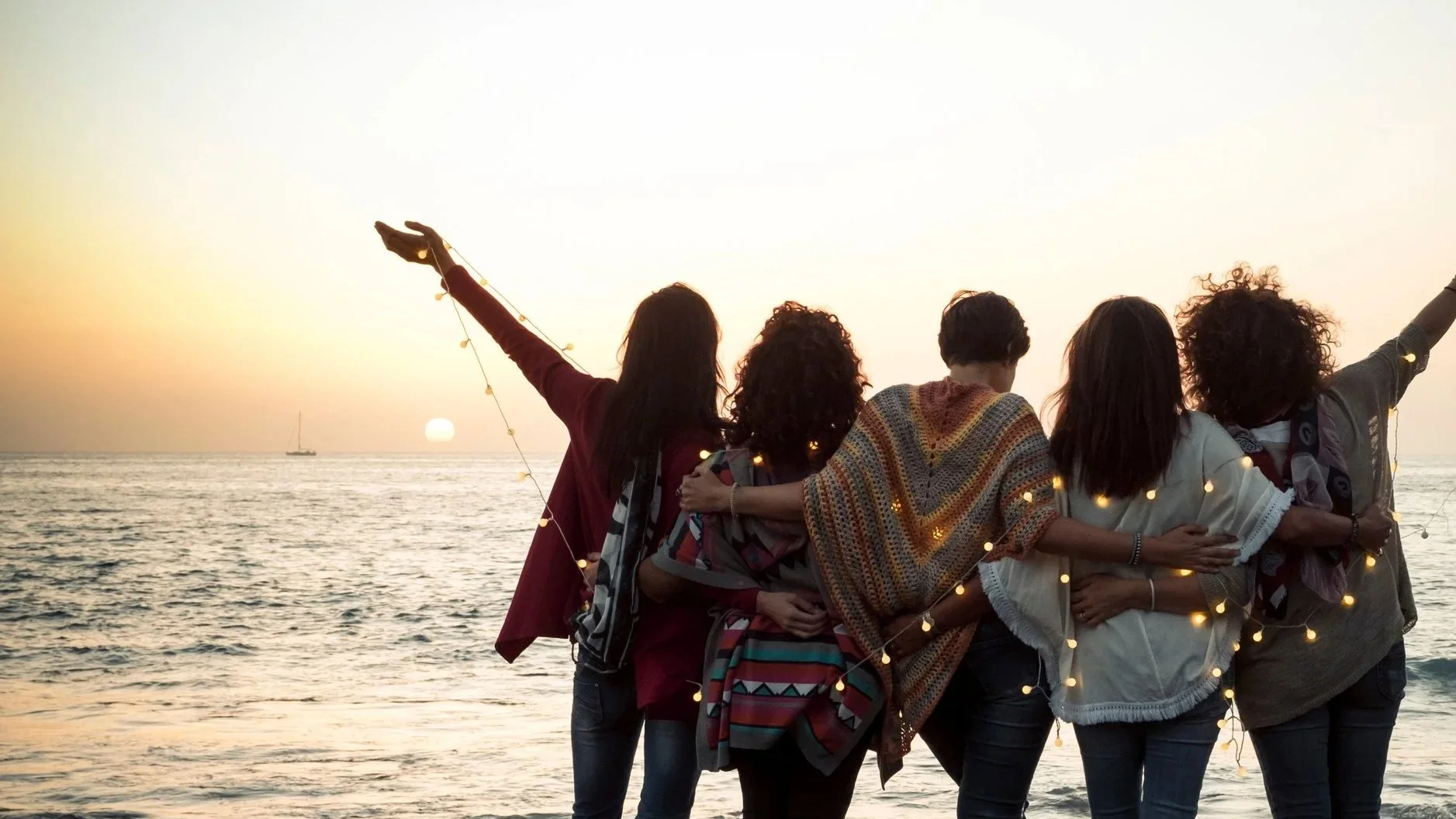 Group of friends at the beach during sunset, embracing and celebrating with string lights.