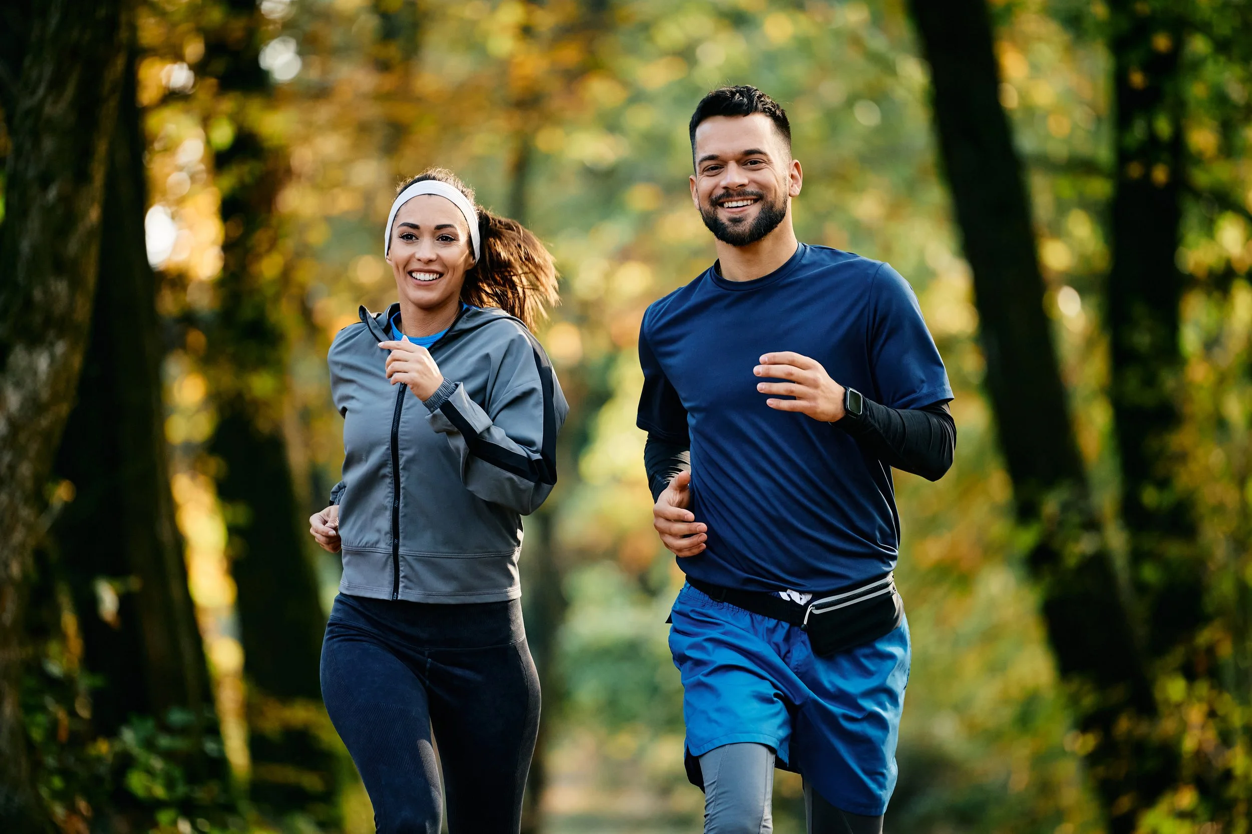 A man and woman jogging together in a forest with green and autumn foliage, both smiling.