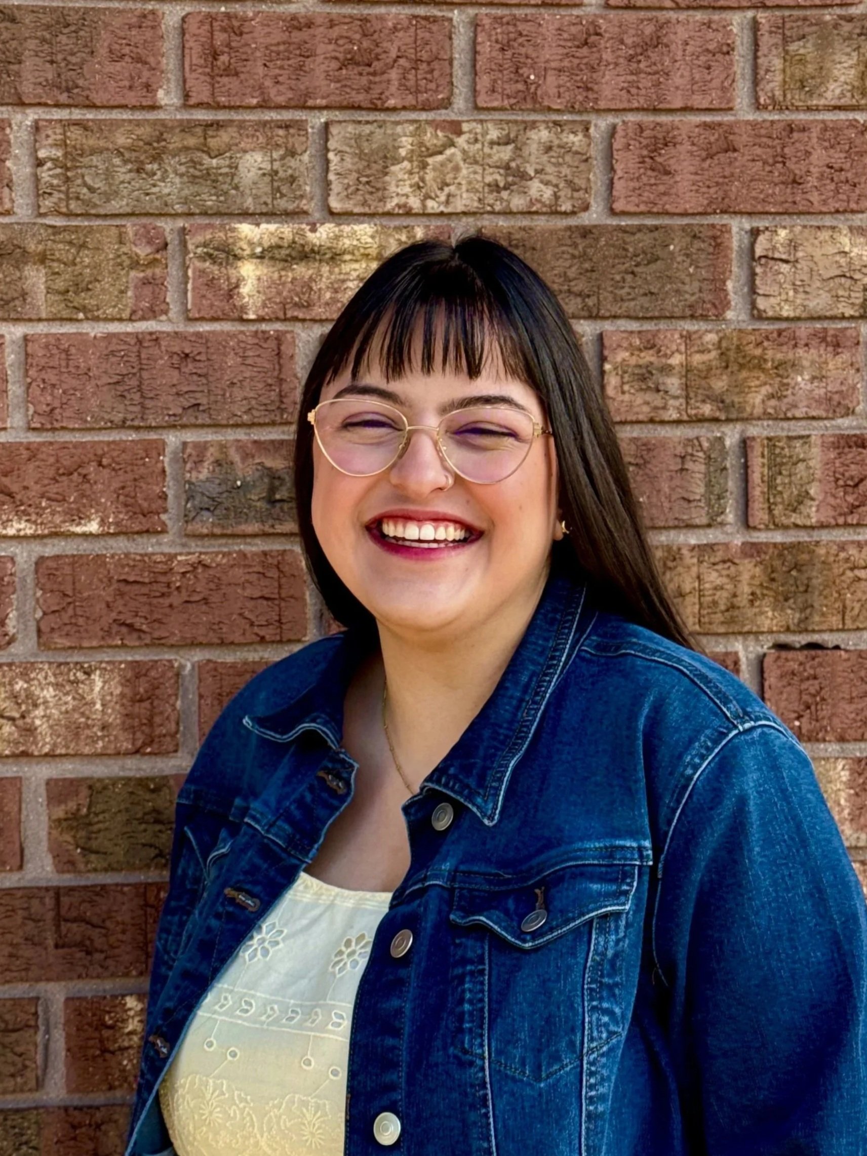 A woman with dark hair, glasses, and a nose piercing, smiling, standing against a brick wall.