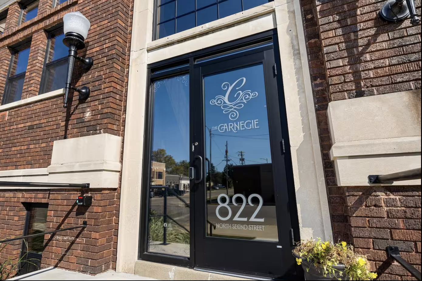 The entrance to The Carnegie, a building on North Second Street with a glass door displaying the name and address, located at 822 North Second Street. The building has brick walls, a black lamp post, and a small garden with yellow flowers near the do