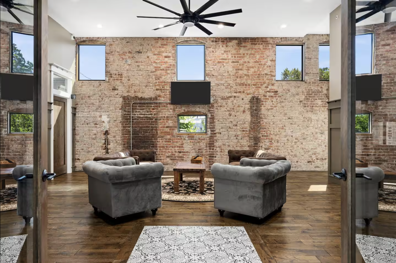 A modern, spacious lounge area with exposed brick walls, multiple windows, and a high ceiling with a black ceiling fan. There are four plush armchairs arranged in a circle around a wooden coffee table, and two large flat-screen TVs mounted on the wal