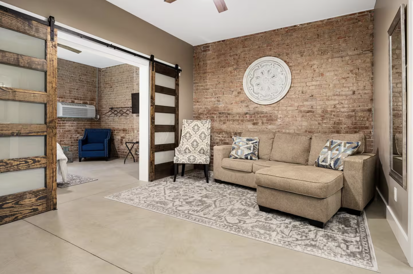 Living room with exposed brick wall, a beige sofa with patterned pillows, an armchair with a floral pattern, a decorative wall piece, and a sliding barn door leading to a bedroom containing a blue armchair, an air conditioning unit, and a small table