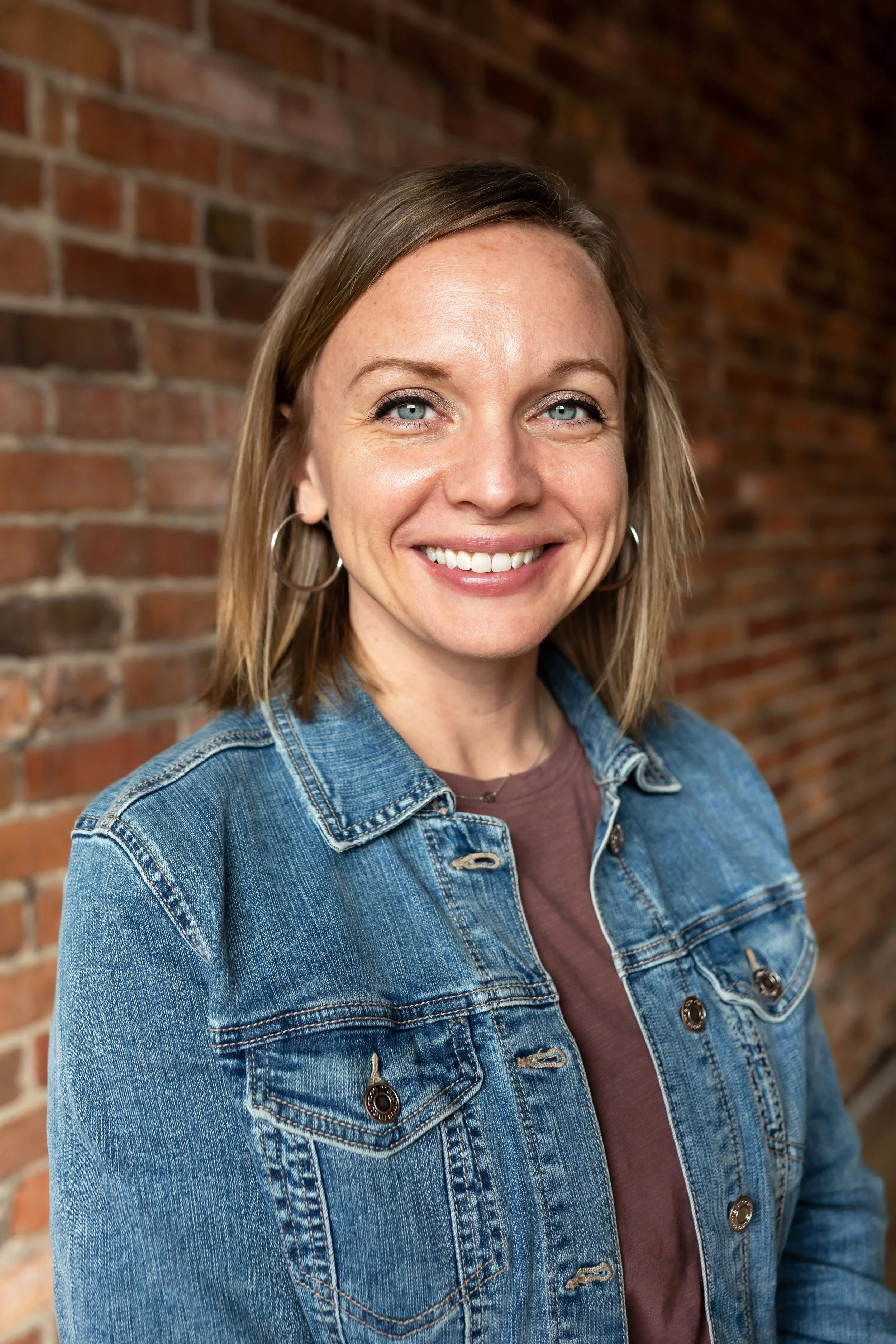 A portrait of a smiling woman with medium, straight strawberry blonde hair, wearing a checked blazer, a burgundy top, and a butterfly-shaped necklace, standing outdoors in front of a brick wall.