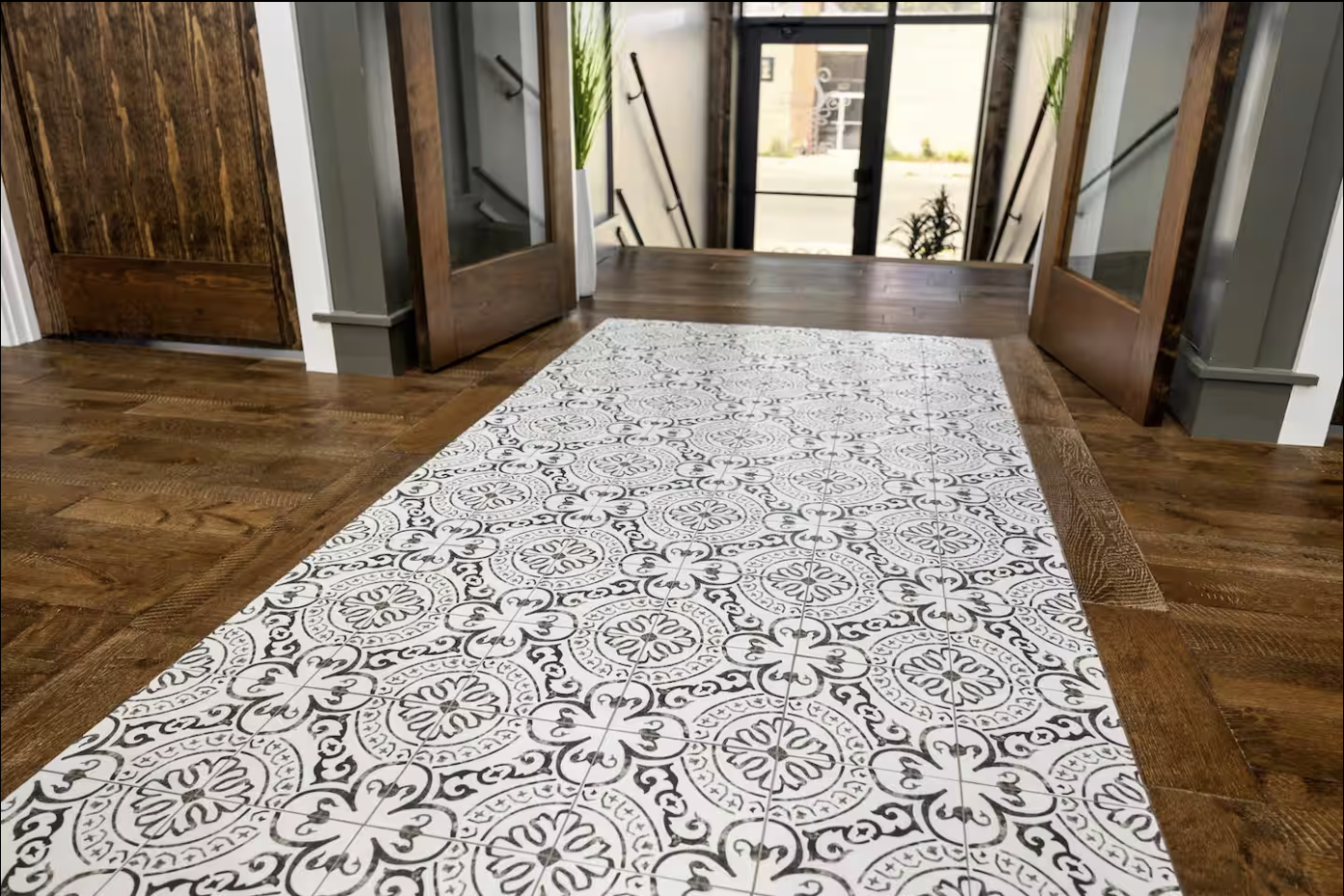 Decorative black and white patterned tile floor strip in a wooden-floored entryway with glass doors and potted plants.