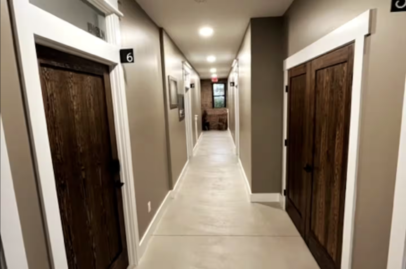 Hallway in an indoor apartment complex with numbered wooden doors, beige walls, white trim, ceiling lights, and a window at the end of the hall.