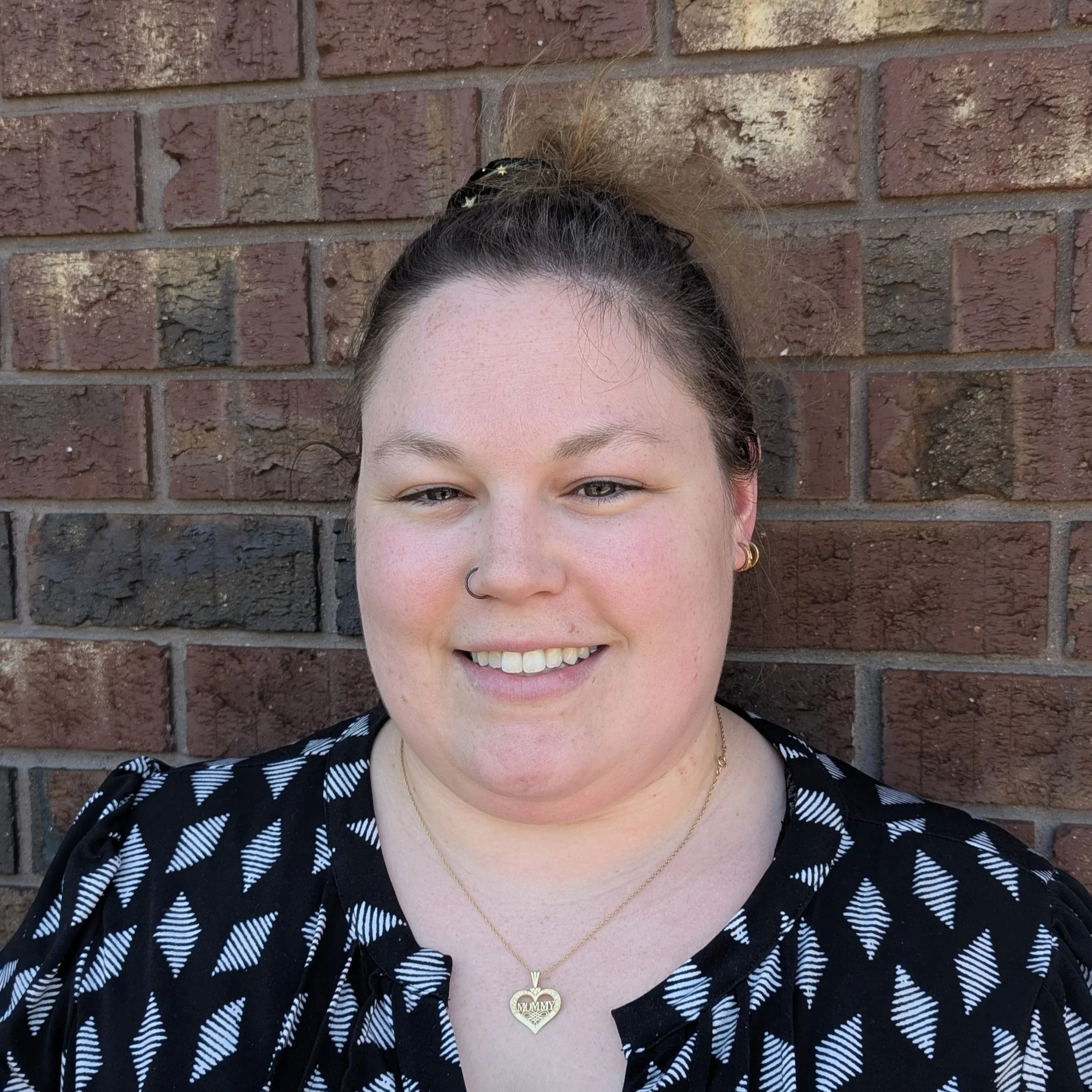 Close-up of a woman's face and upper body standing against a brick wall, smiling, wearing a black and white patterned top, gold jewelry, and a necklace with a heart-shaped pendant.