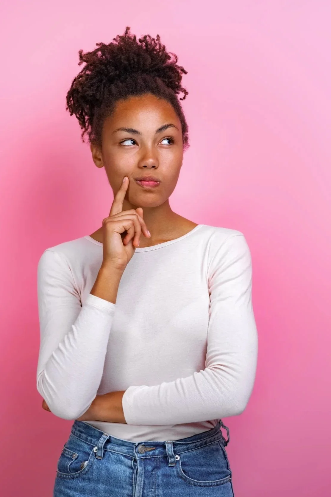 A young woman with curly hair in a bun, wearing a white long-sleeve shirt and jeans, standing against a pink background, touching her cheek thoughtfully.