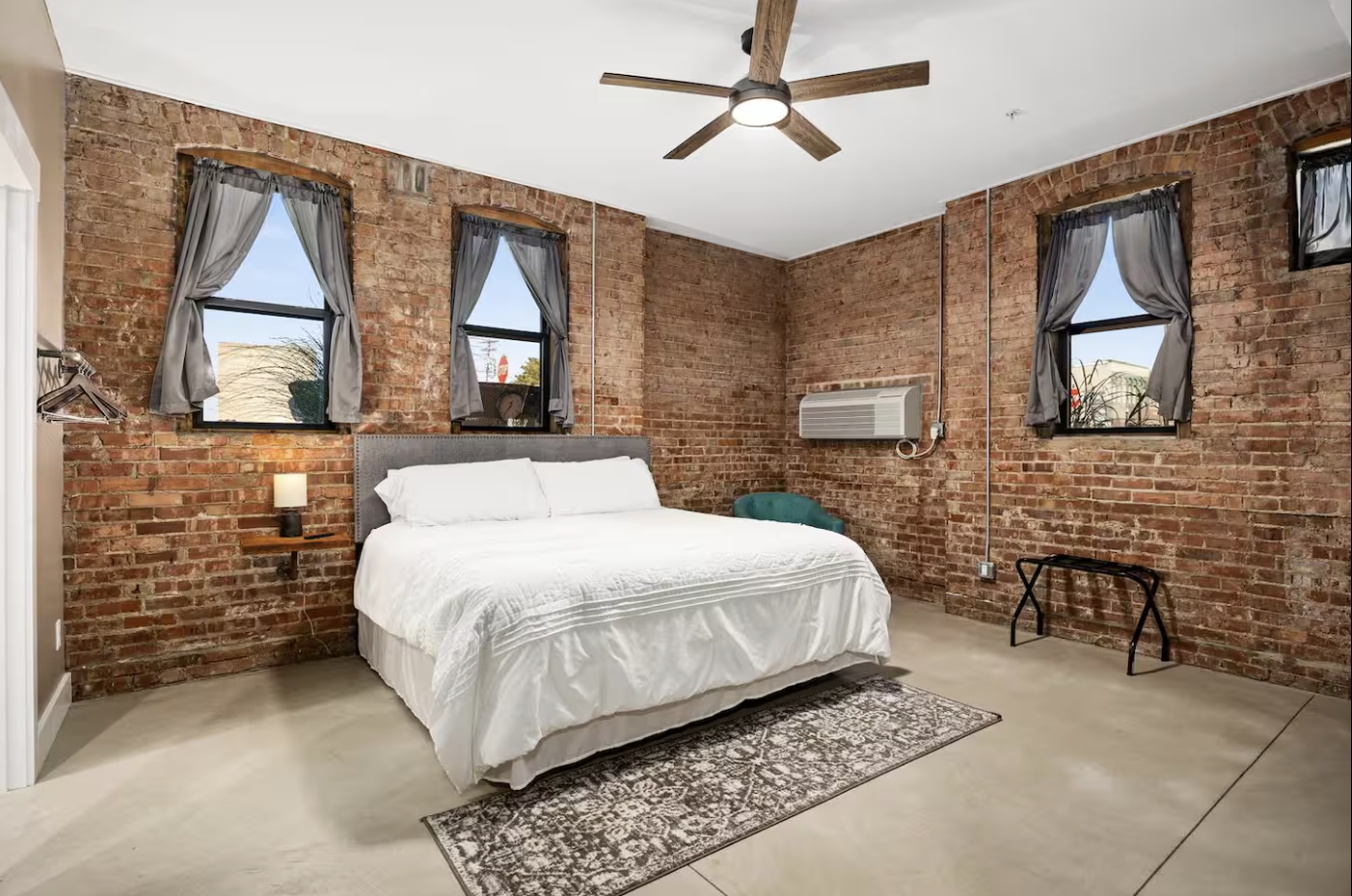 A bedroom with exposed brick walls, a bed with white bedding, three windows with gray curtains, a ceiling fan, a wall-mounted air conditioner, a small side table with a lamp, a green chair, a black luggage rack, and a decorative rug.