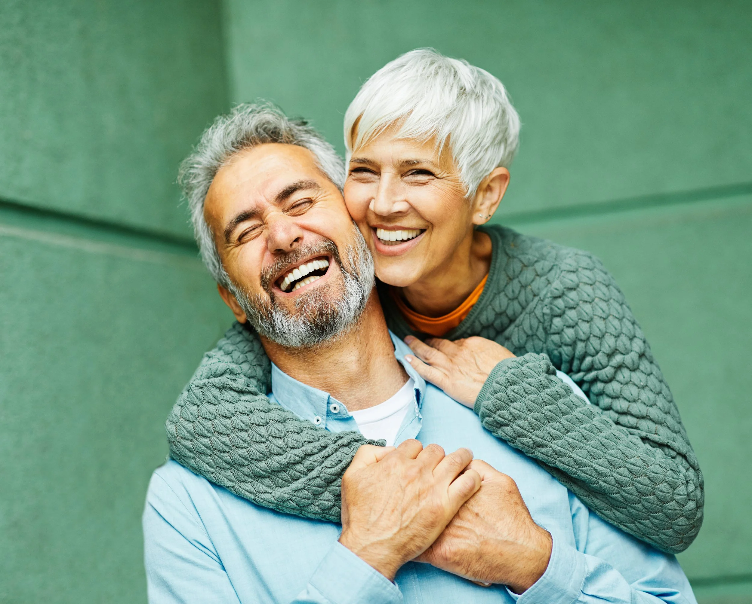 A joyful elderly couple with gray hair, smiling and laughing together, embracing outdoors against a green wall.