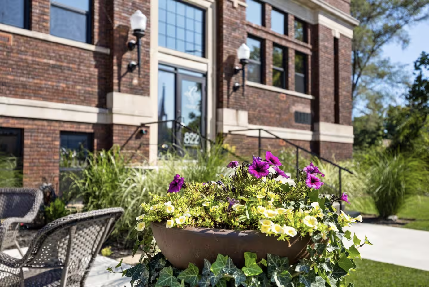 Flower pot with purple, yellow, and white flowers outside a brick building with large windows and a black metal railing